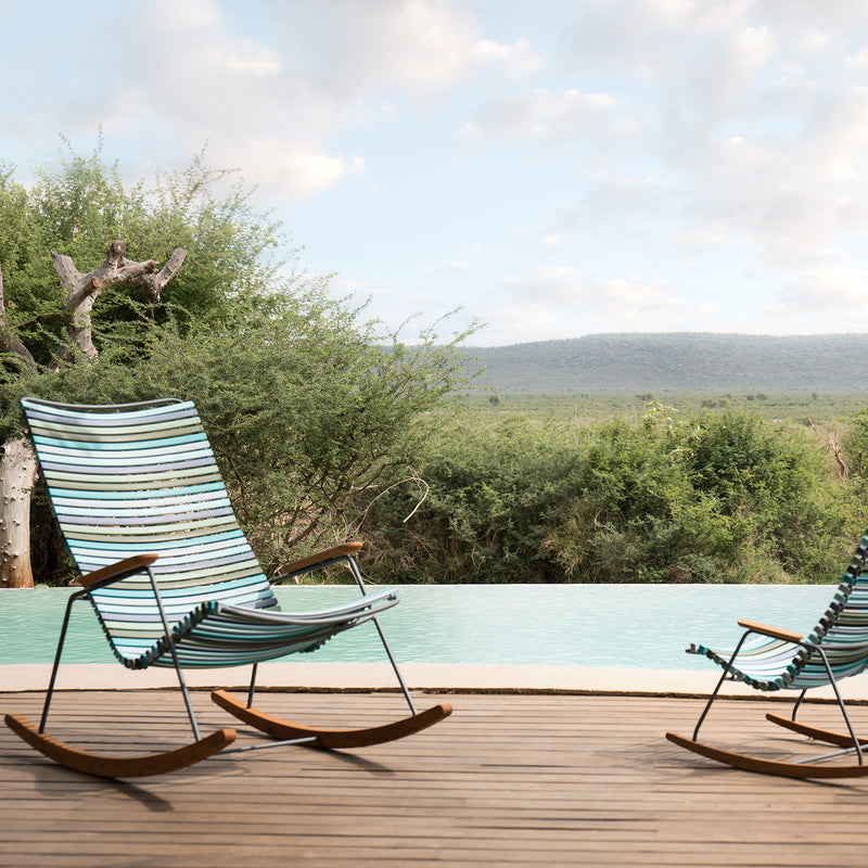 Two Click Rocking Chairs by Houe, made with powder-coated steel and plastic lamellas, adorn a wooden deck beside an infinity pool. Lush greenery stretches behind the pool towards distant hills under a partly cloudy sky, crafting a serene and inviting scene.