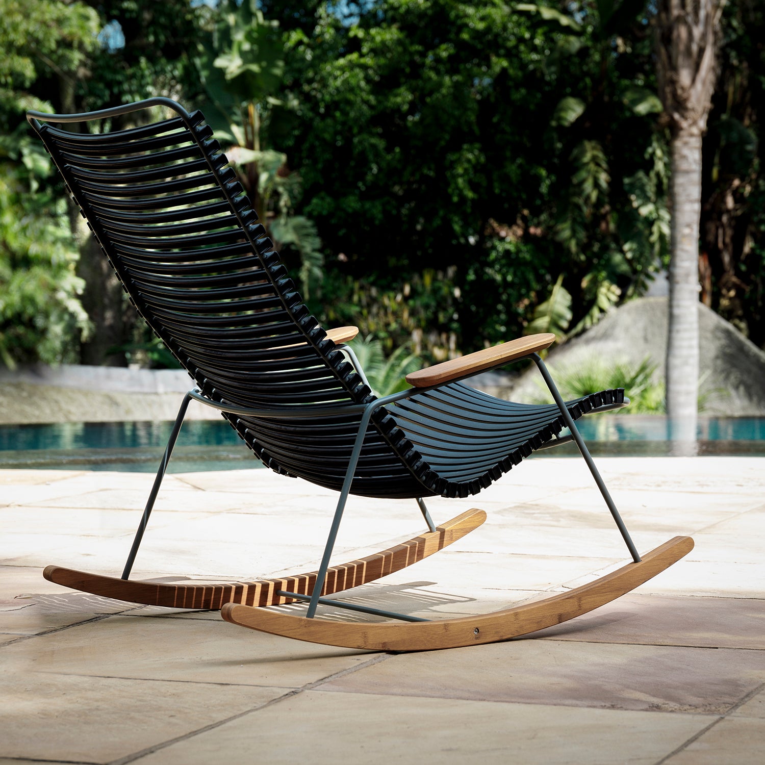A sleek Click Rocking Chair by Houe, with wooden armrests and rockers, sits on a sunlit stone patio. The plastic lamellas complement the serene setting featuring a swimming pool surrounded by lush greenery and trees.