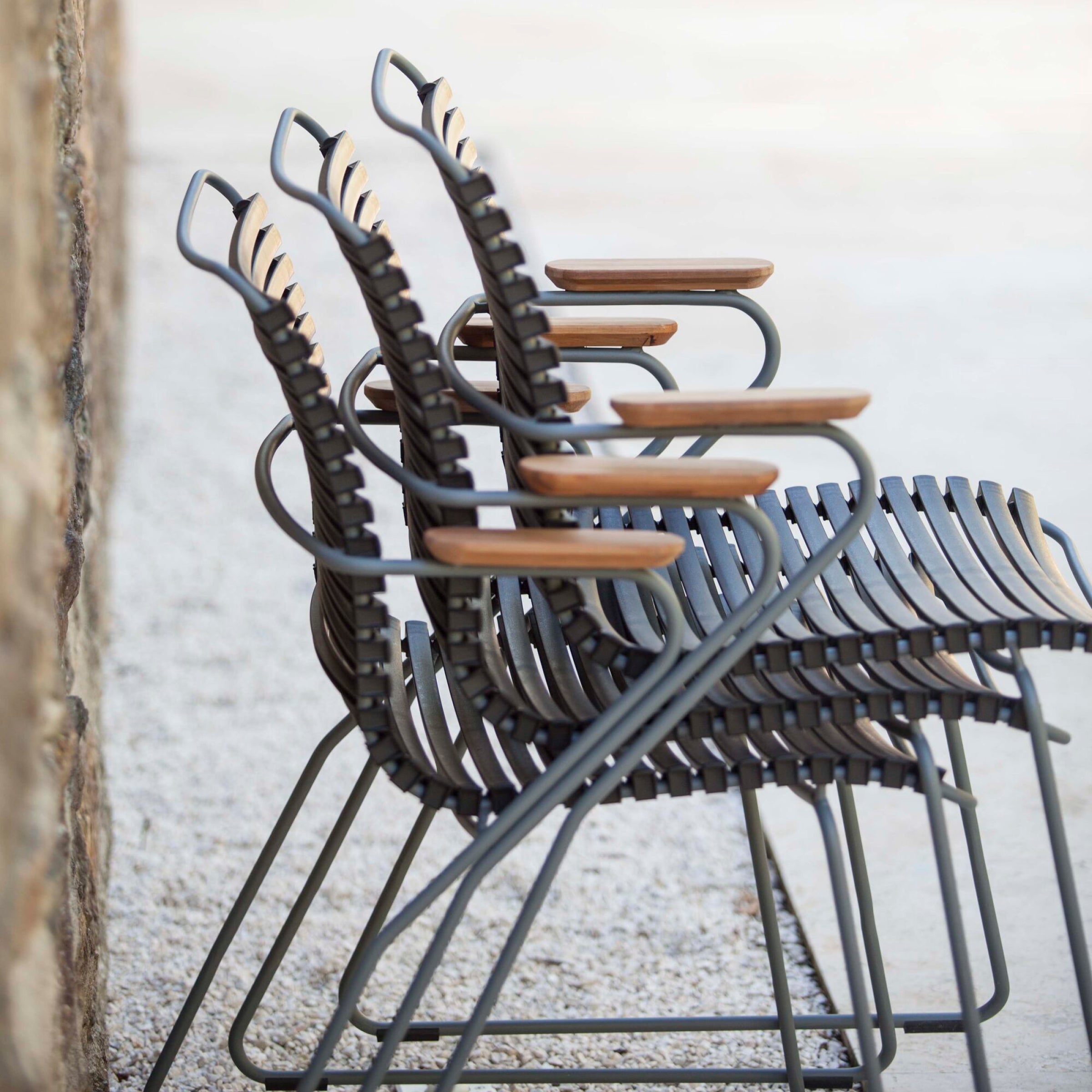 Four Houe Click Dining Chairs with Armrests, featuring weather-resistant slatted seats and wooden armrests, are lined up on a gravel surface next to a stone wall. Their sleek, modern design is highlighted by their angled positioning.