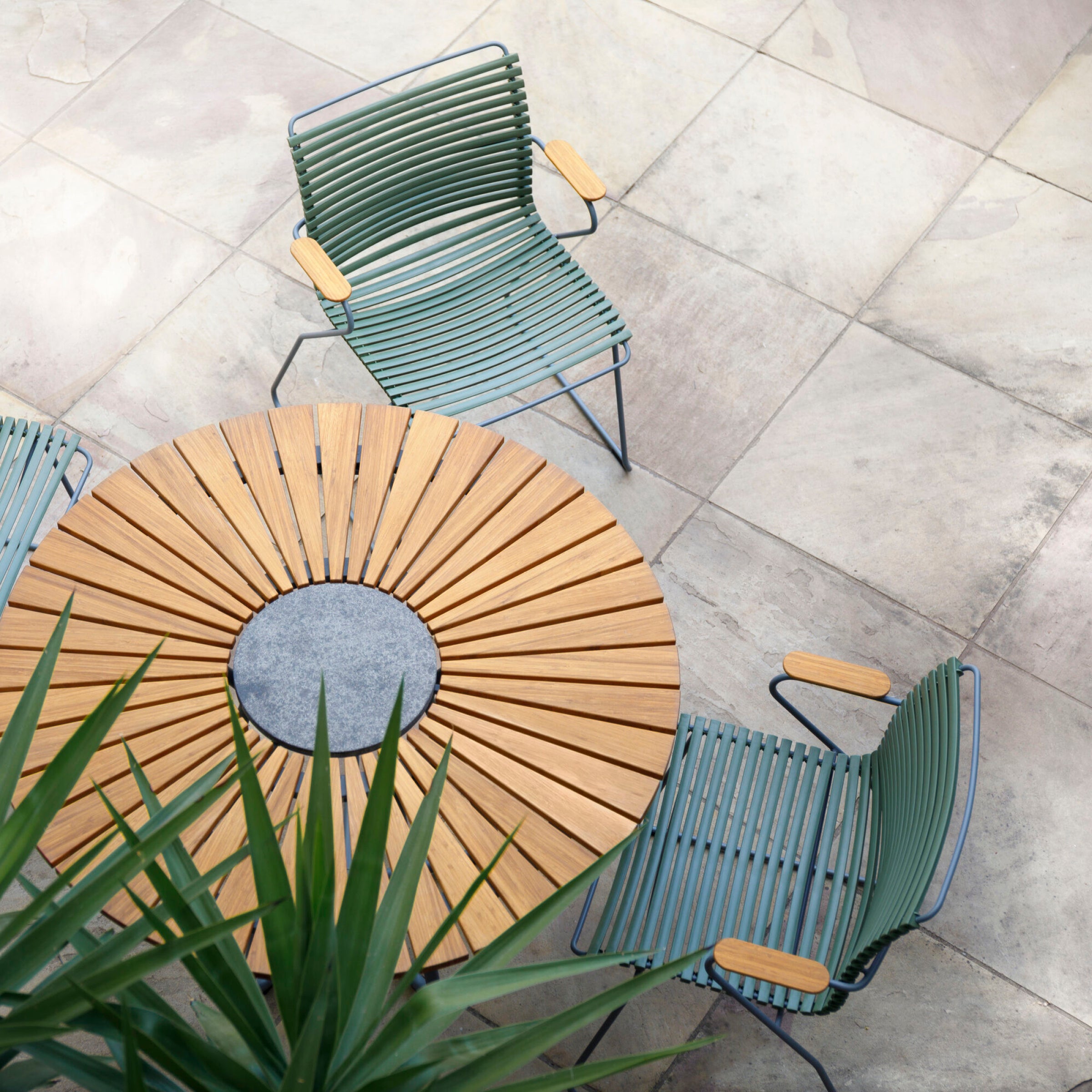 A round wooden table with a sunburst pattern is surrounded by Houes Click Dining Chairs with Armrests - Tall Back. These weather-resistant, green metal chairs have wooden armrests and sit on a tiled patio. A spiky green plant in the foreground enhances the outdoor setting.