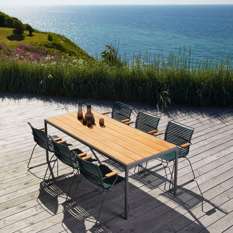 A wooden deck overlooking the ocean features a UV-protected table and eight weather-resistant Houe Click Dining Chairs with Armrest in green, set against grassy hills and a clear blue sky.