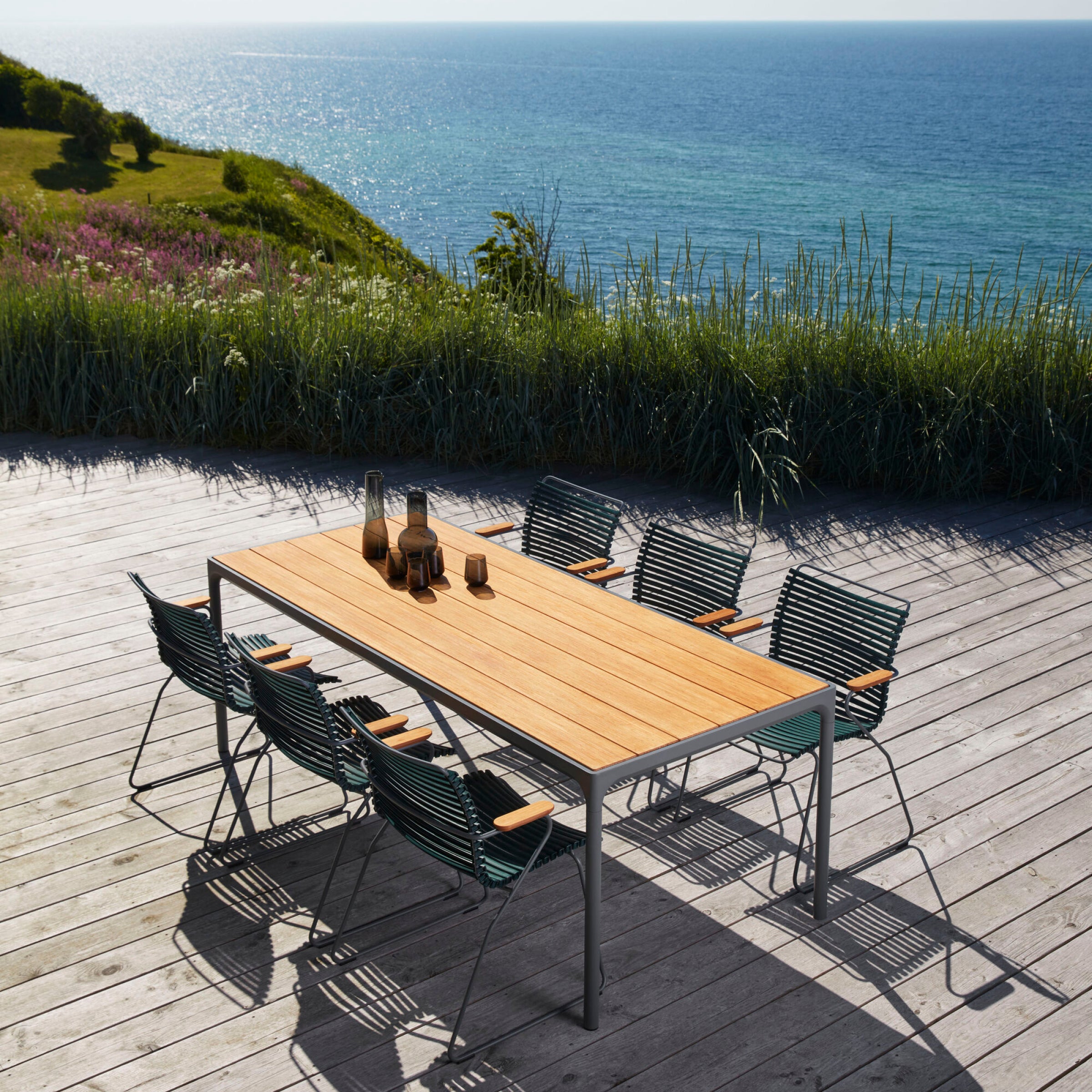 The Click Dining Chair with Armrest from Houe, paired with a wooden table, creates a serene deck setting overlooking the ocean. With grassy hills and a clear sky in view, these UV-protected chairs ensure lasting beauty and tranquility.