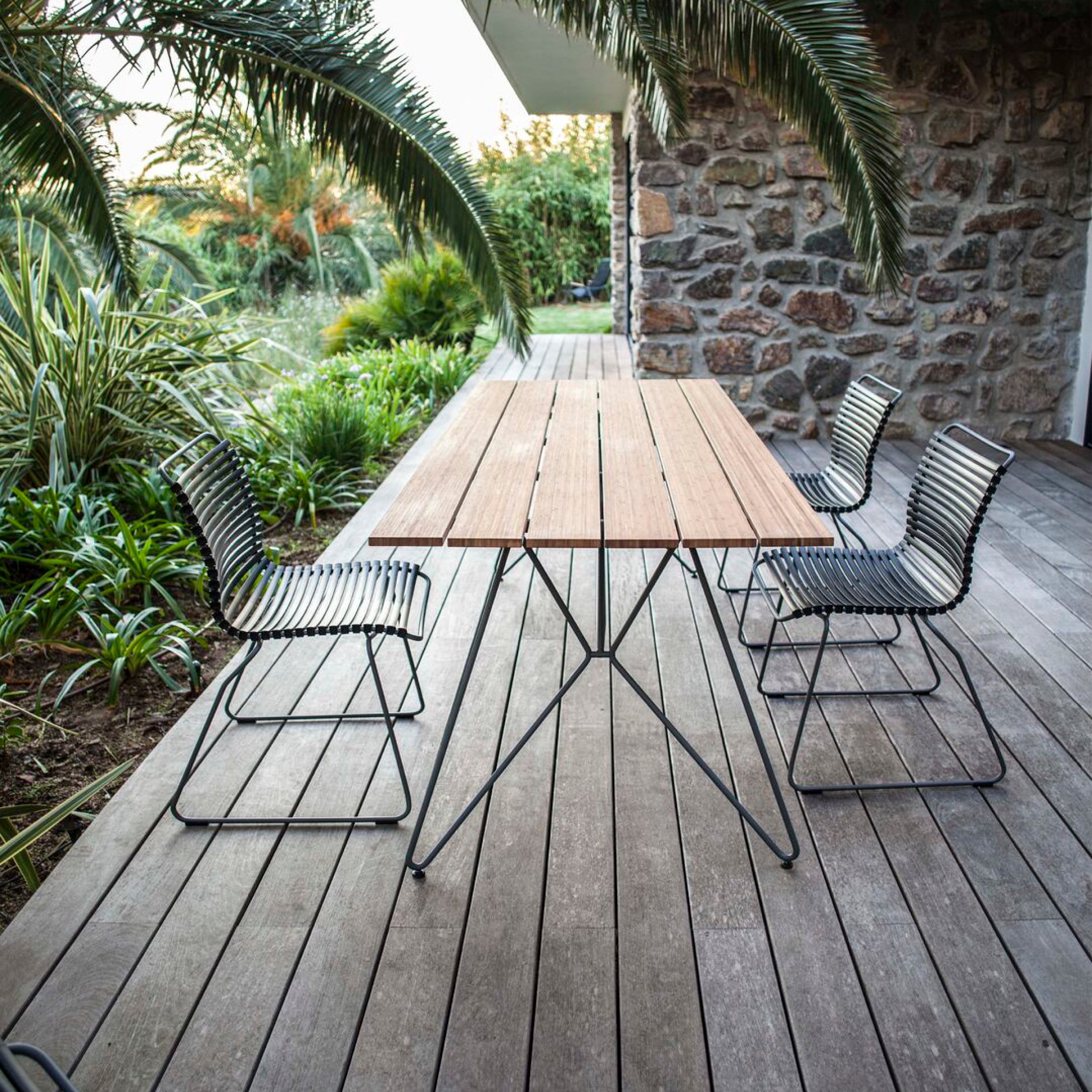 A Houe Click Dining Chair without Armrest set, featuring four weather-resistant metal chairs with plastic lamellas around a wooden patio table, is placed on a wooden deck. Surrounded by lush greenery and palm trees, a stone wall in the background suggests a tropical outdoors.