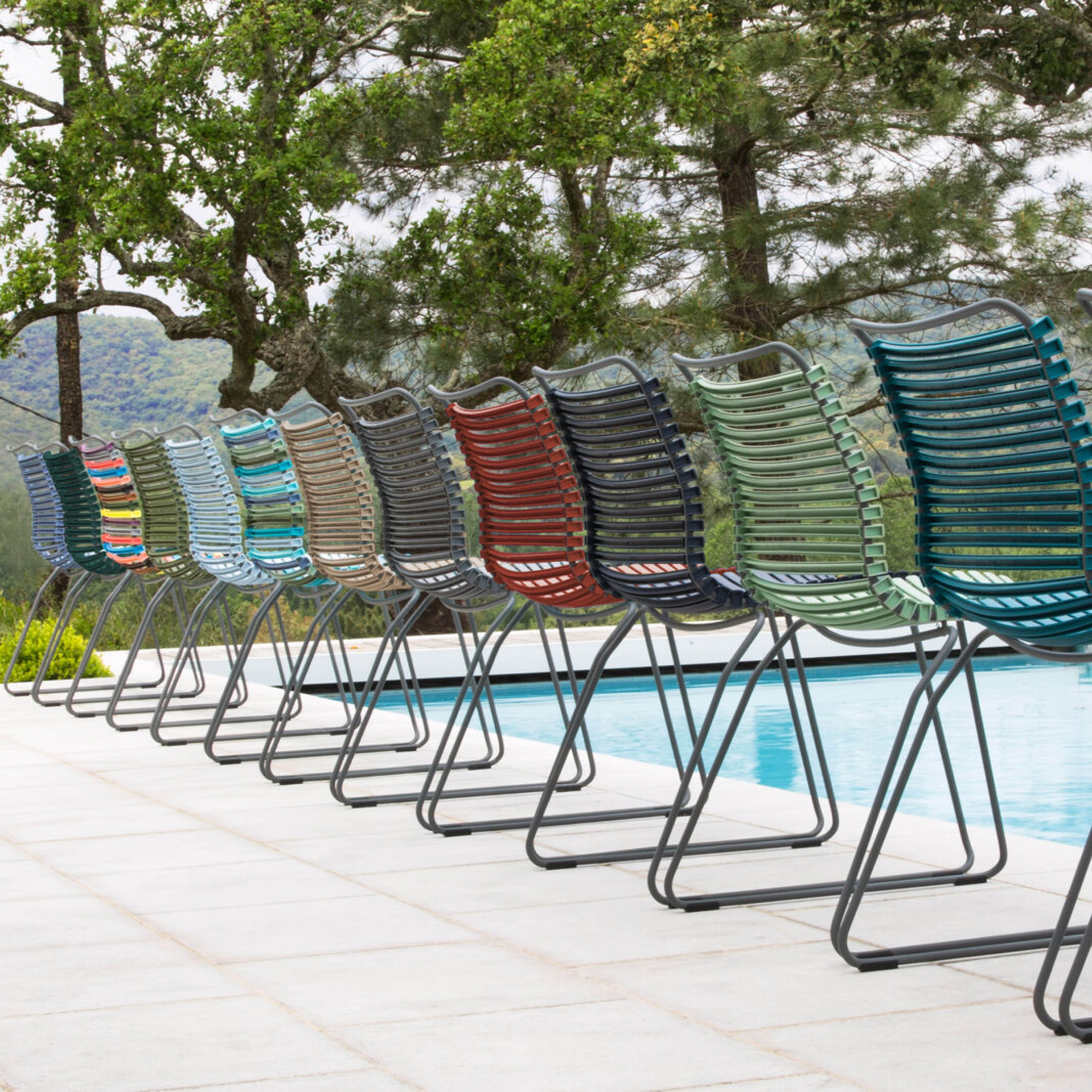 A row of Houes colorful Click Dining Chairs without armrests, featuring metal frames and plastic lamellas in shades like blue, red, green, and gray, sits beside a swimming pool against a scenic backdrop of trees and mountains.