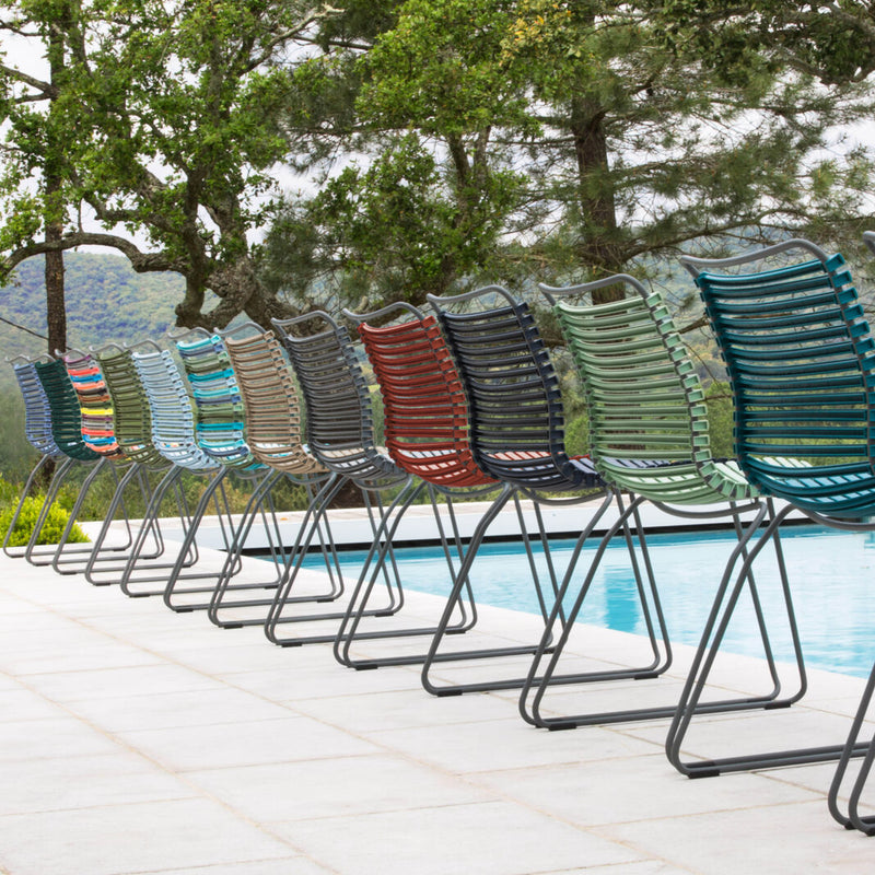 A row of Houes colorful Click Dining Chairs without armrests, featuring metal frames and plastic lamellas in shades like blue, red, green, and gray, sits beside a swimming pool against a scenic backdrop of trees and mountains.