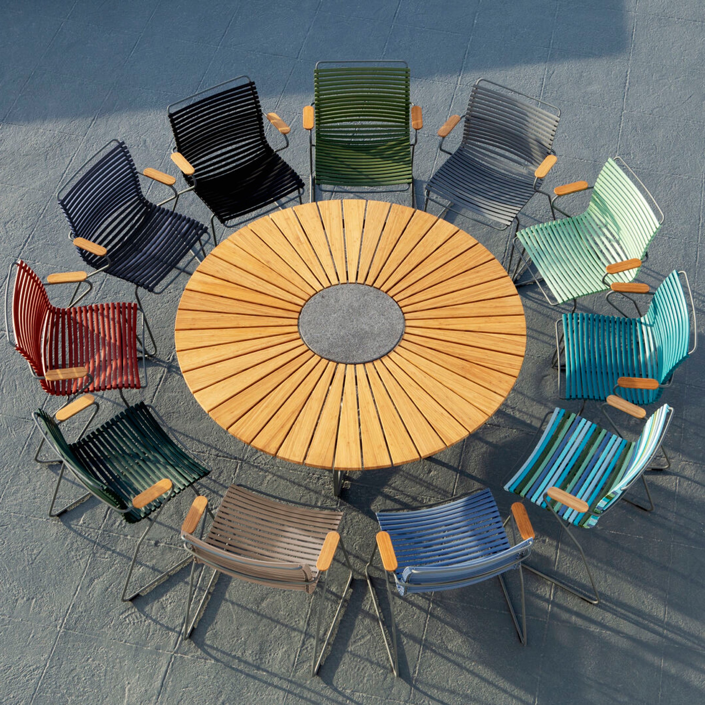 Aerial view of a circular wooden table surrounded by twelve vibrant, weather-resistant outdoor Click Dining Chairs without armrests by Houe, featuring slatted backs, arranged in a circle on a textured gray surface.