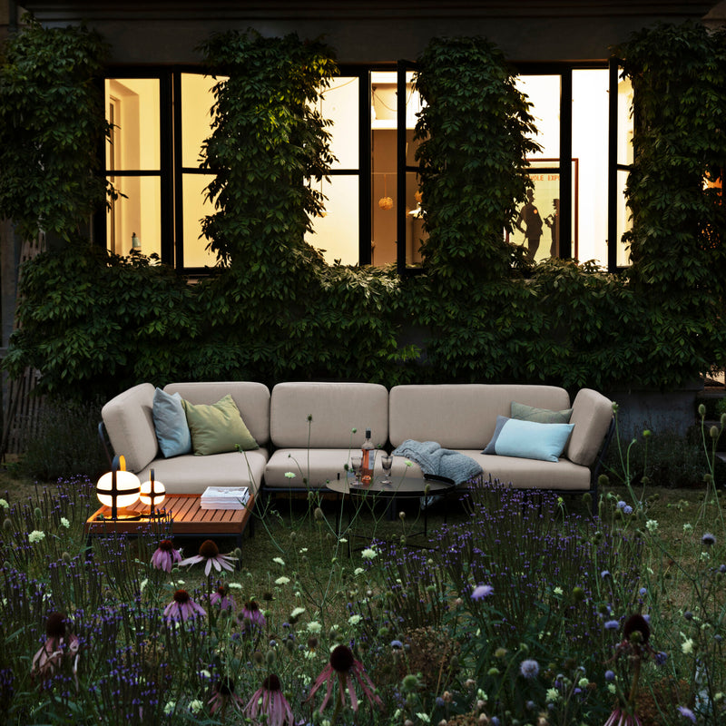 A cozy outdoor seating area features a beige sectional sofa with green and blue cushions. Two lit table lamps illuminate the Level Coffee Table by Houe amidst lush greenery and colorful flowers, while powder-coated aluminum accents enhance the illuminated windows in the background.