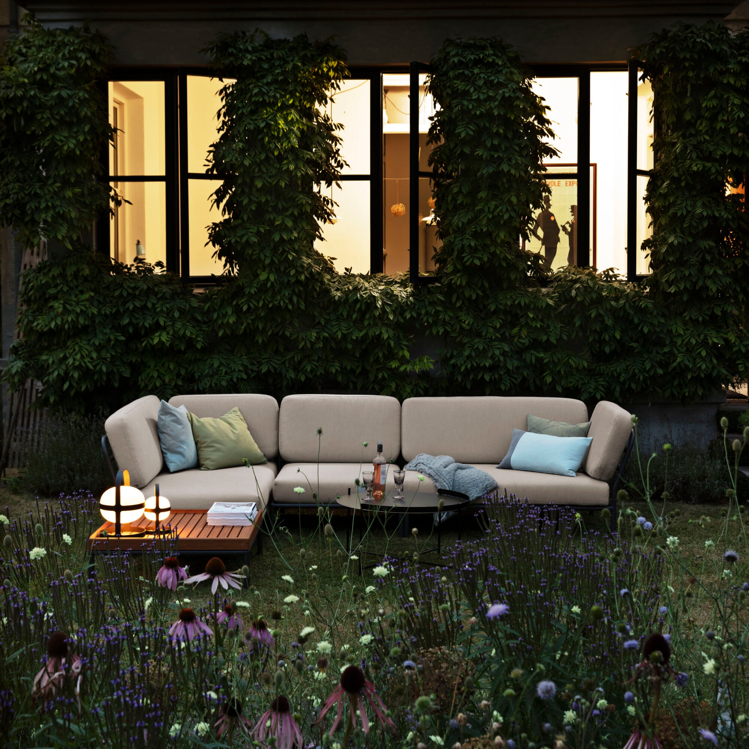 An inviting outdoor retreat showcases Houes Level Corner Sofa in a cozy beige Sunbrella fabric, decorated with plush cushions. A wooden table holds glowing lamps and drinks, surrounded by lush greenery and facing tall, illuminated building windows.