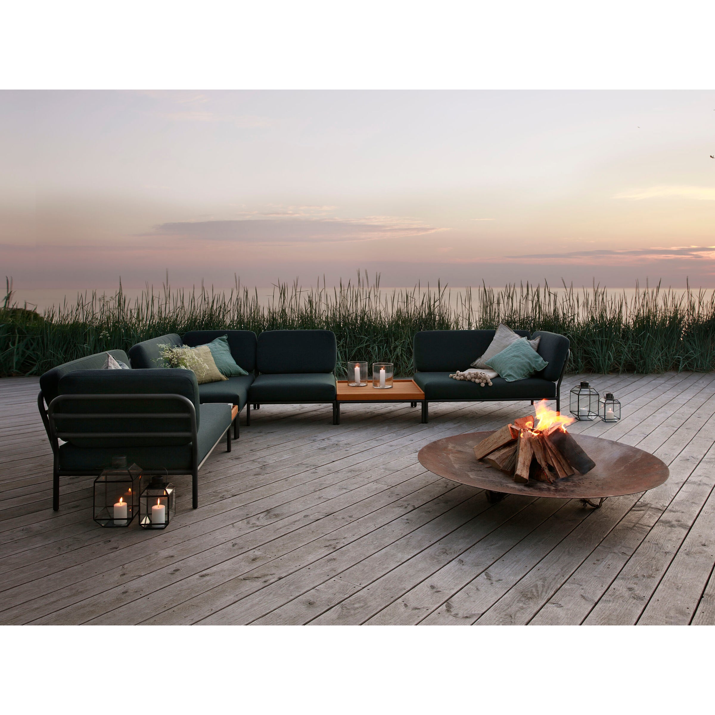 An outdoor seating area includes dark green sofas with beige cushions on a wooden deck. A round fire pit radiates warmth, surrounded by Houes Level Coffeetable and bamboo tables. Soft candle lanterns glow nearby, framed by tall grass and a sunset over tranquil water.