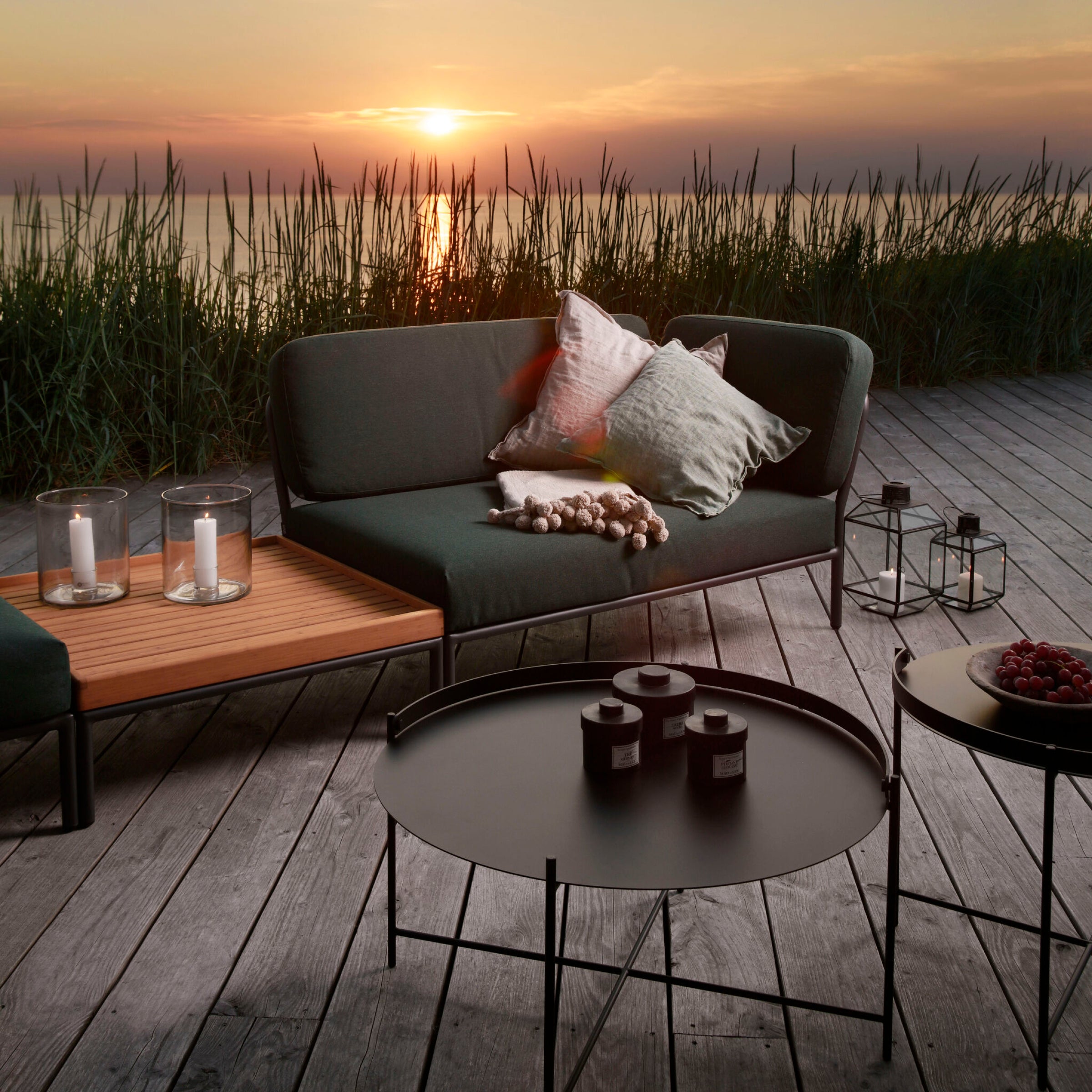 The outdoor wooden deck showcases Houes Level Corner Sofa with Sunbrella fabric cushions. Candles, lanterns, and a tray table with cherries enhance the ambiance as tall grasses by a serene lake mirror the sunsets warm glow.
