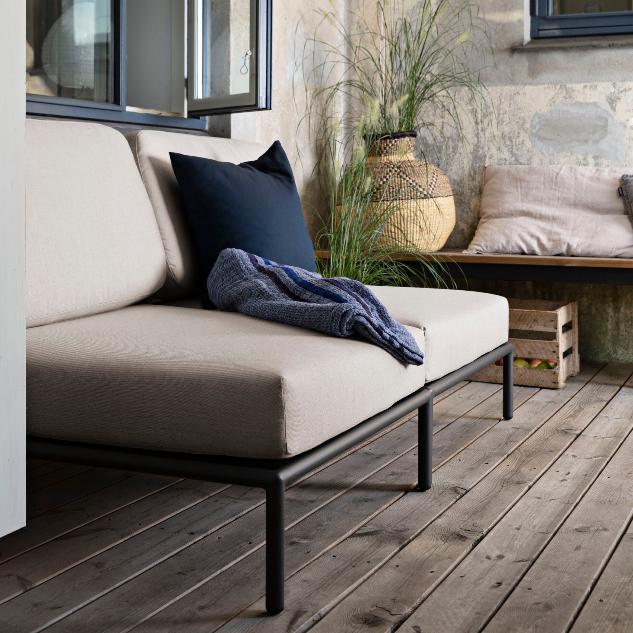 A modern patio showcases the Level Lounge Chair by Houe, with light cushions on a powder-coated aluminum frame. A dark pillow and blue blanket adorn the seat. A wicker basket with a plant and a wooden bench with pillows enhance the textured wall ambiance.