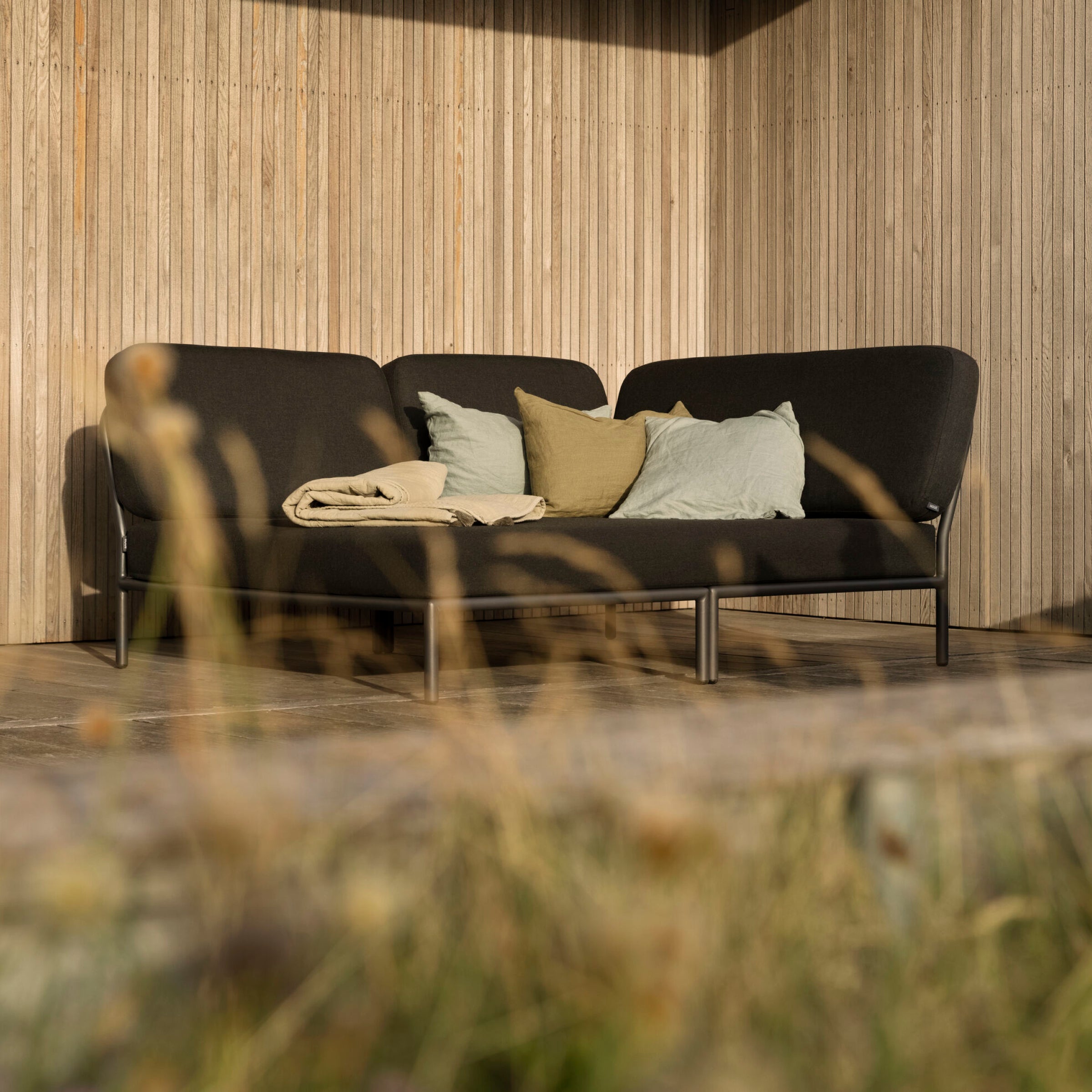 A modern patio features the Houe Level Cozy Corner, a sleek black sectional with LEVEL modules. Its adorned with water-repellent green and beige pillows, complemented by a beige blanket. Wooden slat walls and blurred foreground grasses enhance the serene, natural atmosphere.