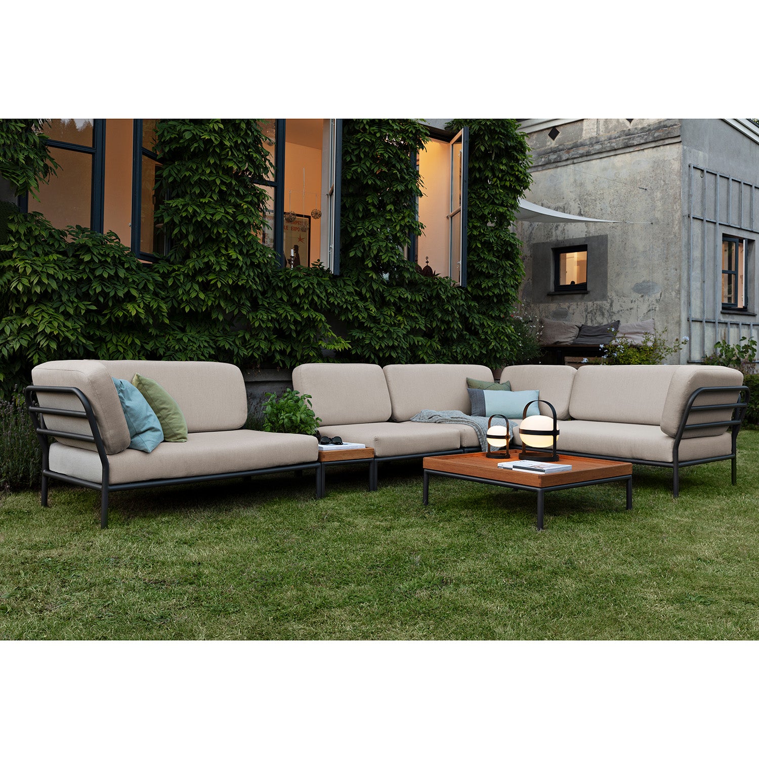 The outdoor seating area showcases the Level Corner Sofa by Houe with cream cushions on powder-coated aluminum frames, elegantly set on the grass. A low wooden table with two lanterns is central. In the background, a building features vine-covered walls and large windows, adding to the serenity.