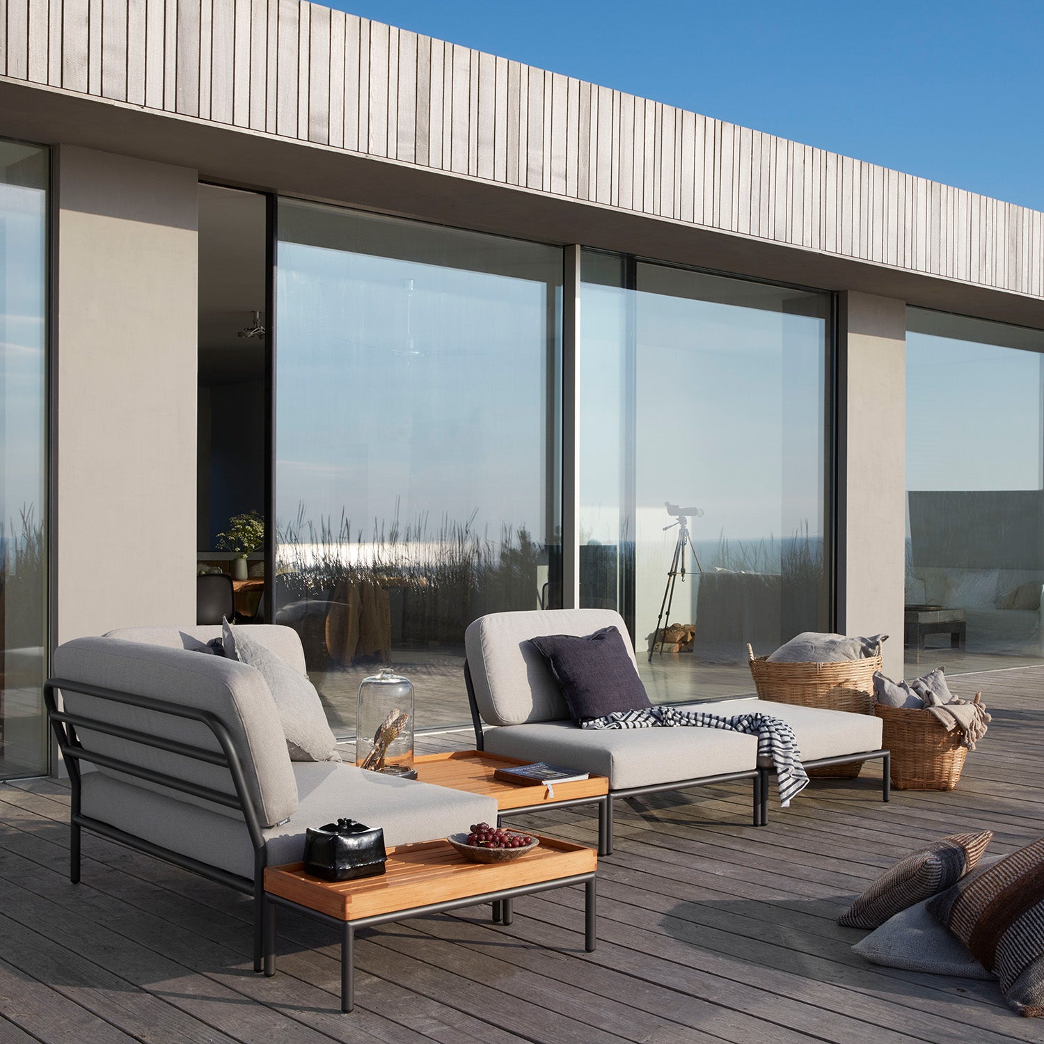 An outdoor patio features two gray cushioned lounge chairs beside a Houe Level Coffeetable, topped with a glass pitcher and bowl of grapes. Large glass doors reflect the blue sky. A woven basket and telescope sit in the background, accentuated by sleek powder-coated aluminum designs.