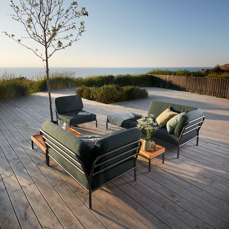 The outdoor area showcases Houes LEVEL modules, including two sofas, two Level Lounge Chairs, and a low table on a wooden deck. A small tree adds charm as the ocean sparkles beneath a blue sky. The furniture features Sunbrella fabric for lasting style.