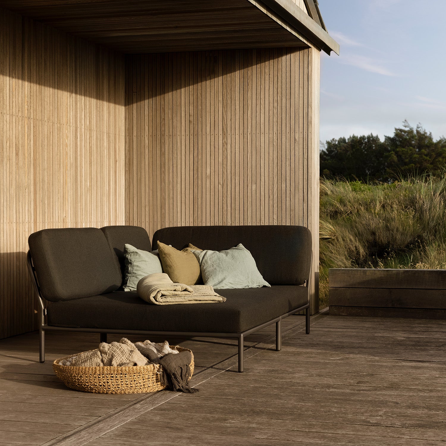 The patio scene showcases the Houe Level Cozy Corner, a dark cushioned sofa with water-repellent fabric and assorted pillows. A wicker basket with blankets rests on the wooden deck next to the modern wooden building, surrounded by greenery under a clear sky.