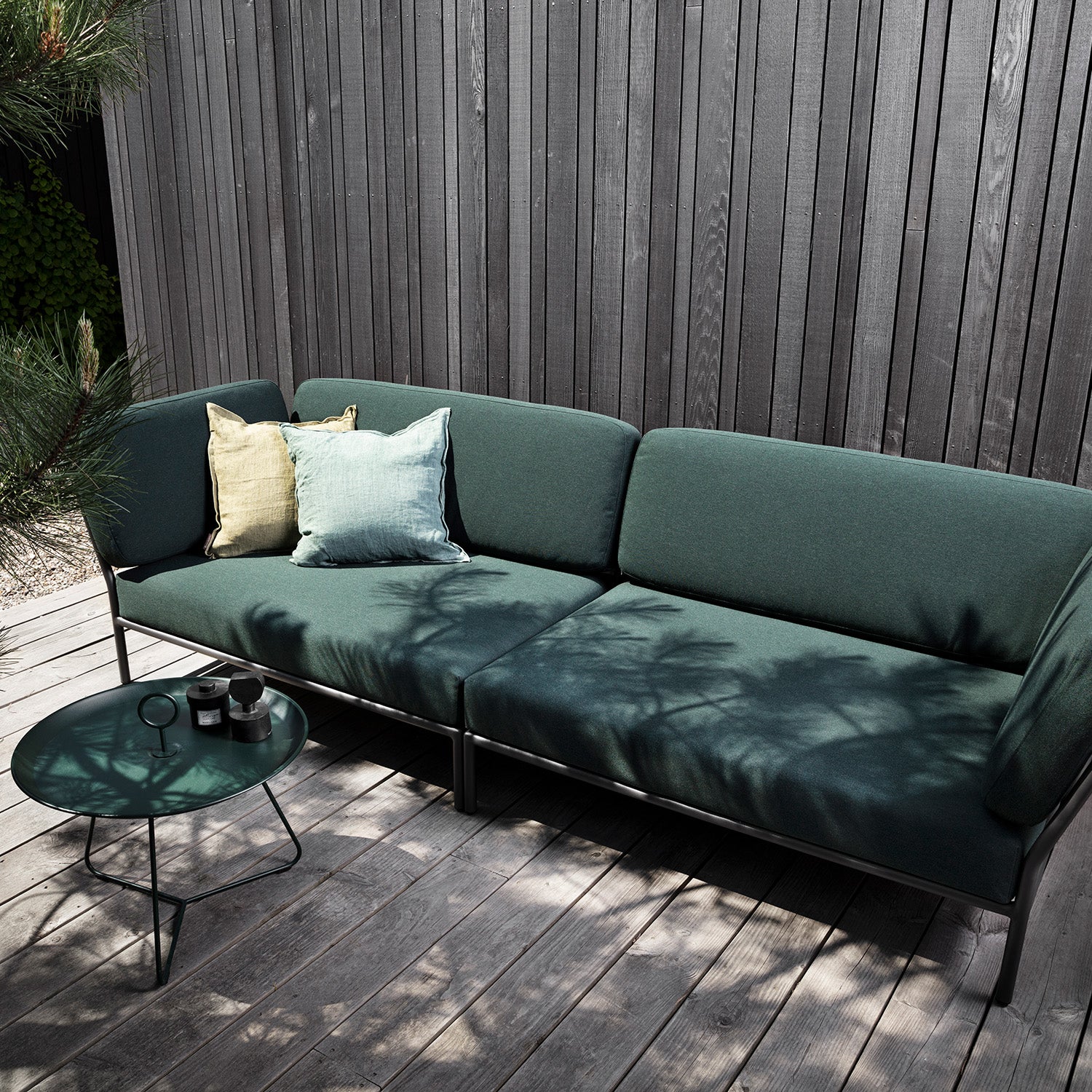 An outdoor area features the Houe Level Corner Sofa with green cushions, decorated with Sunbrella fabric in light yellow and aqua pillows. A round metal powder-coated aluminum table supports two candles, complementing the dark gray wooden fence and wooden deck where tree shadows dance.