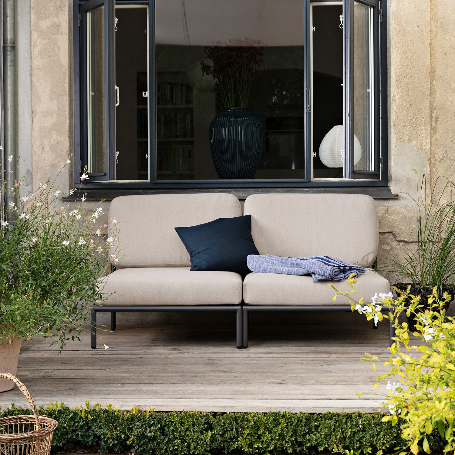 A cozy patio features the Houe Level Lounge Chair with a navy Sunbrella pillow and blue blanket. The chair sits on a wooden deck surrounded by lush greenery, while a large dark-framed window offers views of the homes interior.