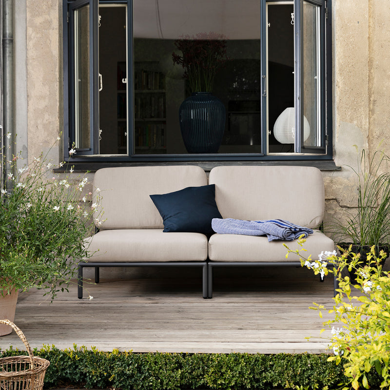 A cozy patio features the Houe Level Lounge Chair with a navy Sunbrella pillow and blue blanket. The chair sits on a wooden deck surrounded by lush greenery, while a large dark-framed window offers views of the homes interior.