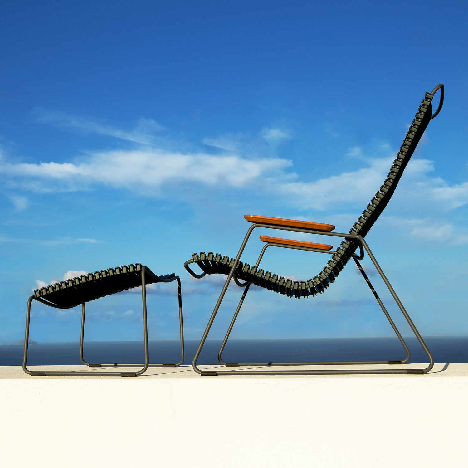 A Click Footrest by Houe sits on a patio, paired with a modern lounge chair under a clear blue sky dotted with clouds. Crafted with UV-protected metal frames and woven seats, the set promises lasting style. The sea shimmers in the background, enhancing this peaceful setting.