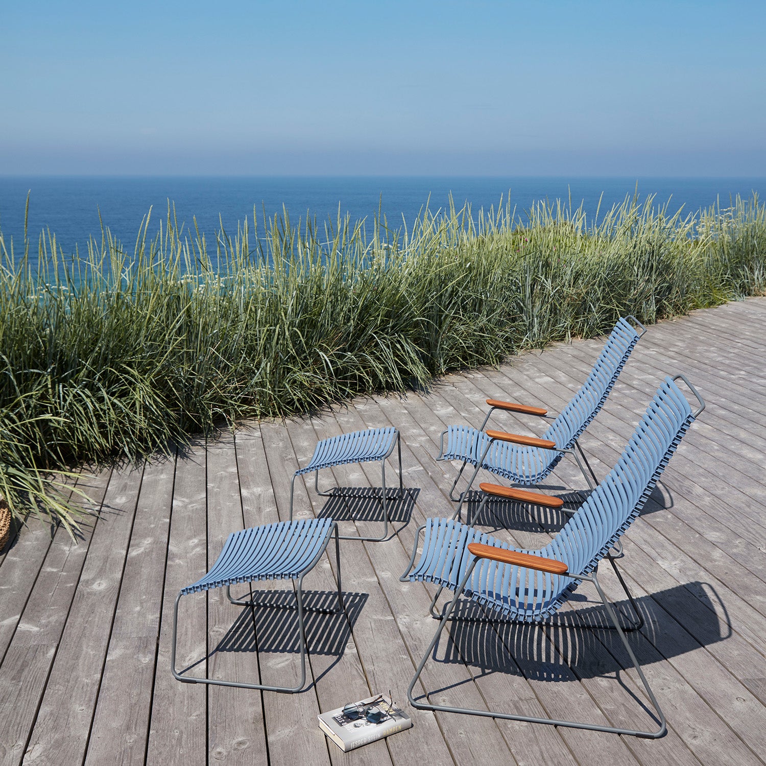 Four blue outdoor chairs with wooden armrests sit on a wooden deck overlooking the ocean, surrounded by tall grass. Beside the UV-protected chairs rests a small notepad and pen. These resilient seats stand strong under the clear blue sky, accompanied by a Houe Click Footrest.