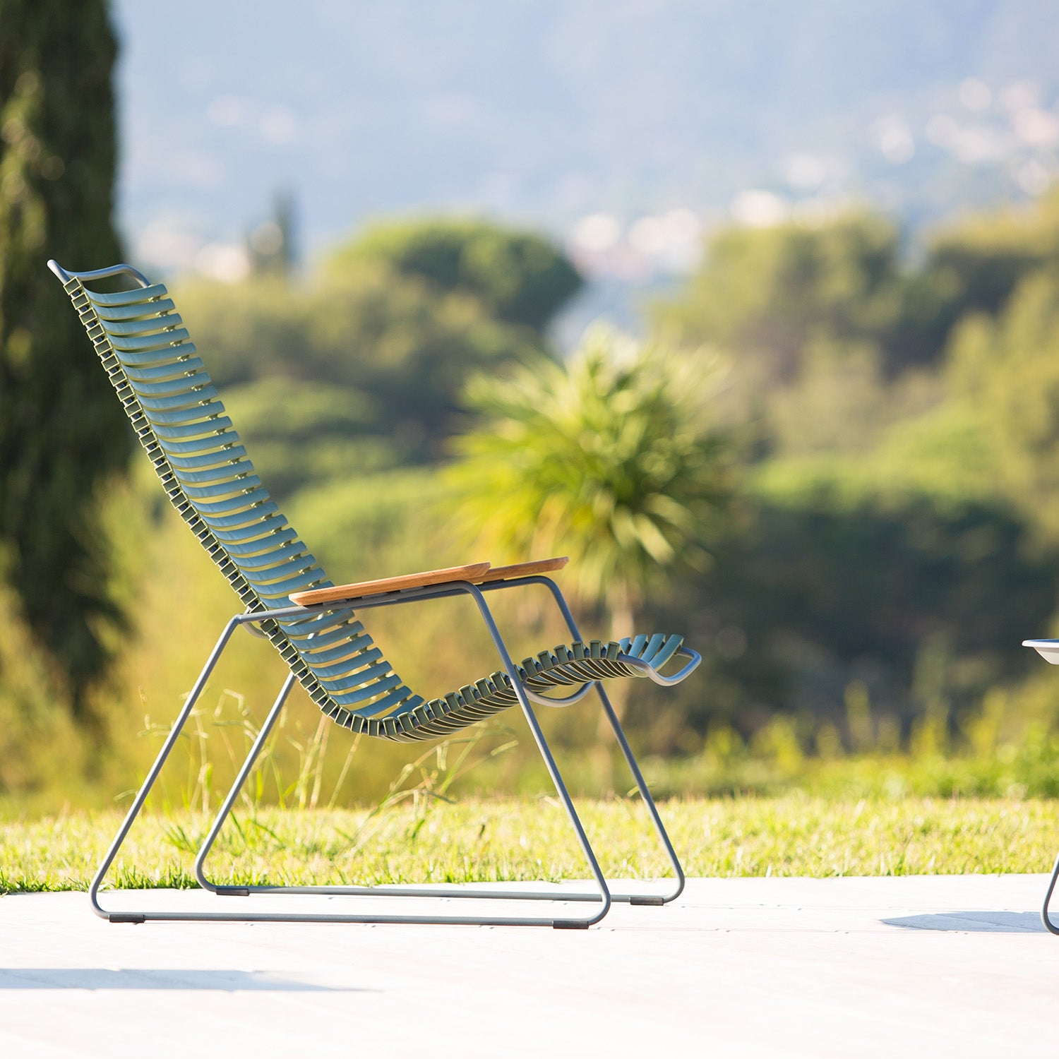 The Click Lounge Chair by Houe, designed for patios, features wooden armrests with plastic lamellas and a metal frame. It stands out against the backdrop of lush greenery and hills under a clear sky.