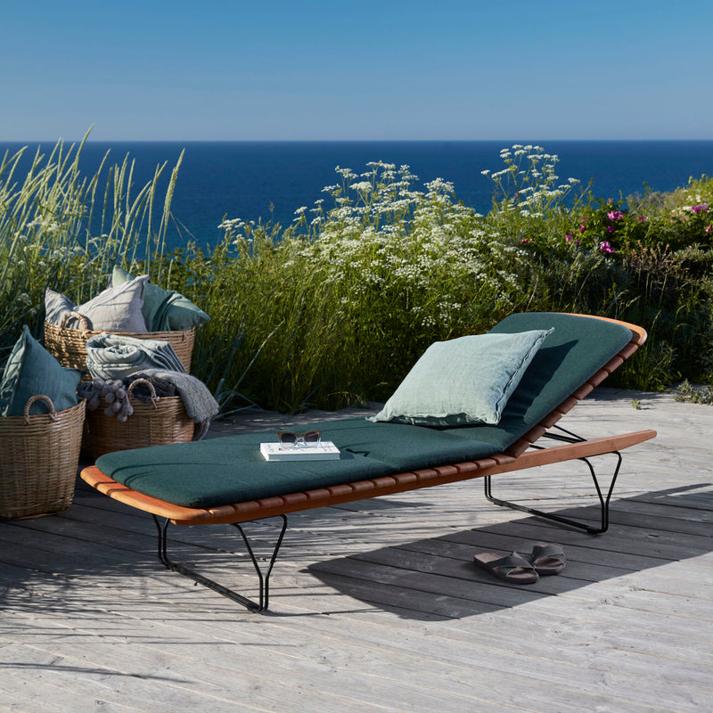 A MOLO Sunbed from Houe, complete with a pillow, graces a wooden deck overlooking the ocean. Its surrounded by greenery and white flowers beneath a blue sky. Baskets of pillows sit against black steel railings, while a book and glasses rest on the bamboo lamellas of the sunbed.