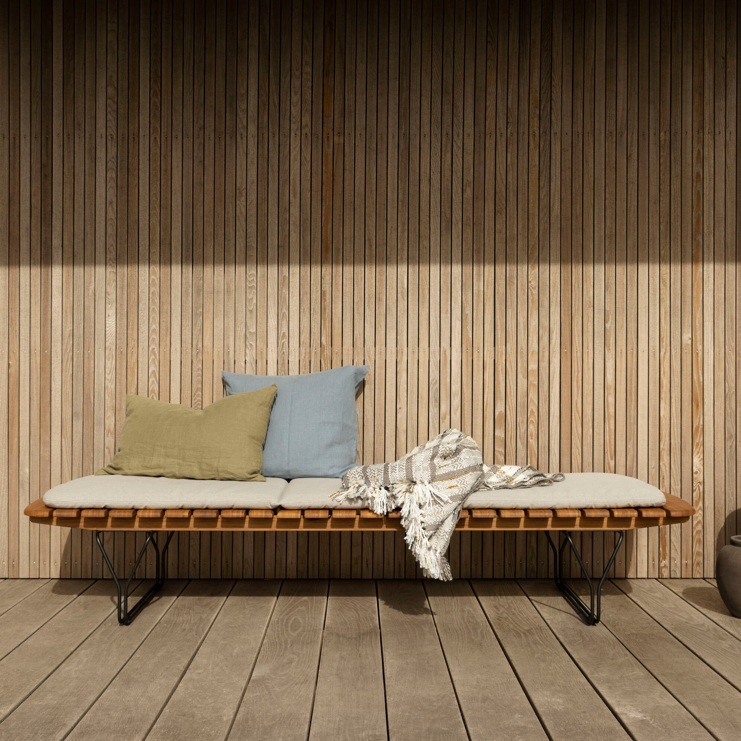 The Houe Molo Sunbed, with a sleek powder-coated black steel frame, features a minimalist slatted wood backrest and light cushion. Complemented by two pillows and a patterned throw against a wood panel wall, it brings modern elegance to your patio.