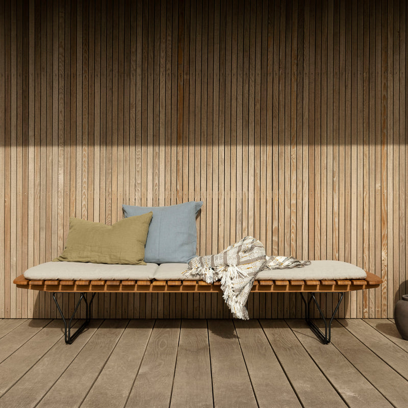 The Houe Molo Sunbed, with a sleek powder-coated black steel frame, features a minimalist slatted wood backrest and light cushion. Complemented by two pillows and a patterned throw against a wood panel wall, it brings modern elegance to your patio.