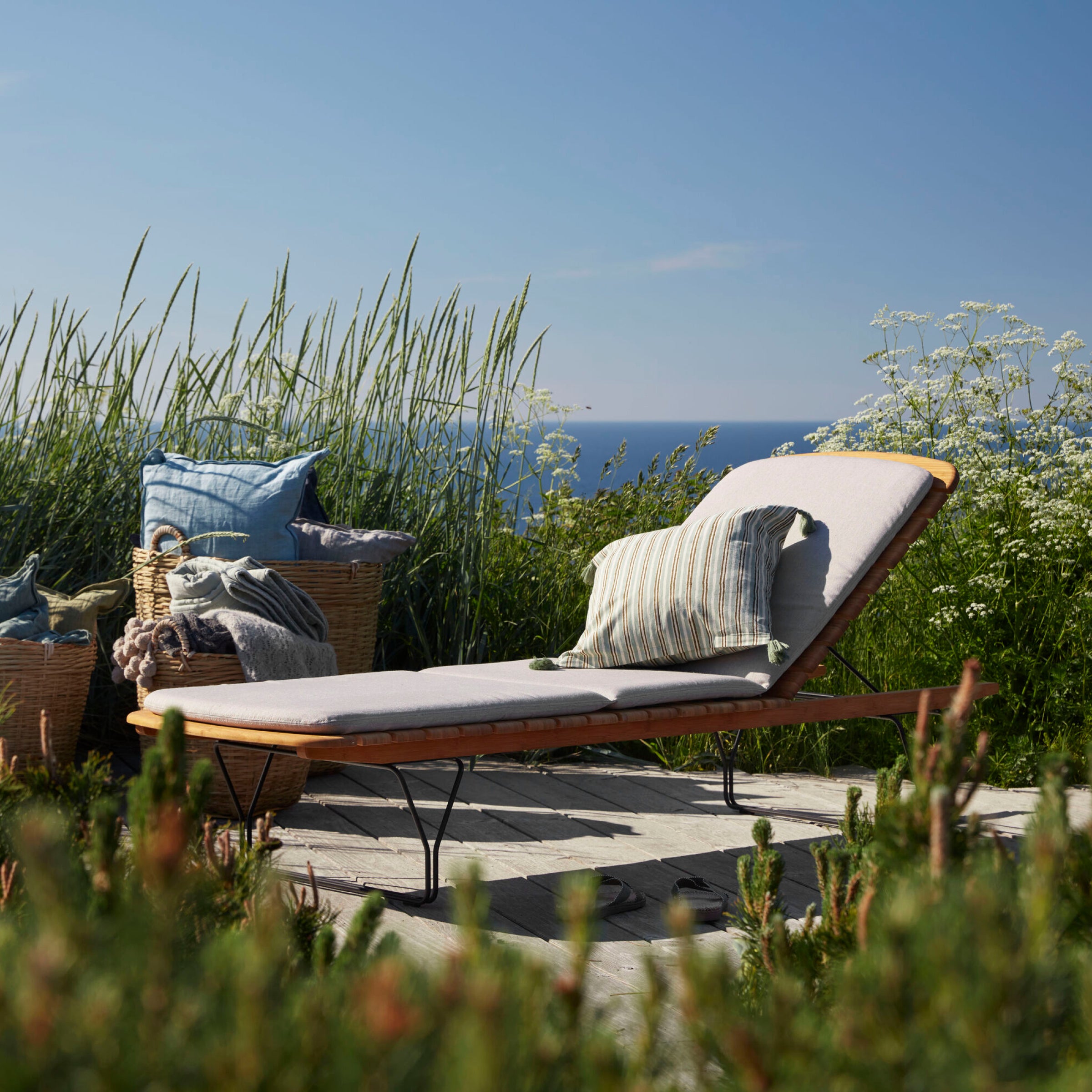 A wooden lounge chair with a cushion and striped pillow sits on a deck amidst tall grass. Nearby, two wicker baskets filled with blue pillows add charm. Under the clear sky, the ocean beckons, flanked by Molo Sunbeds from Houe, elegantly crafted from powder-coated black steel and bamboo lamellas.