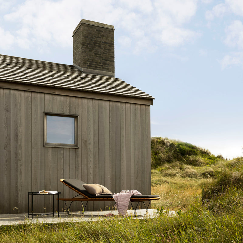 A tranquil wooden cabin with a chimney is set in a grassy landscape. A HOUE Molo Sunbed featuring bamboo lamellas and powder-coated black steel frames supports a book and glass, while a pink blanket drapes over it beneath the partly cloudy sky.