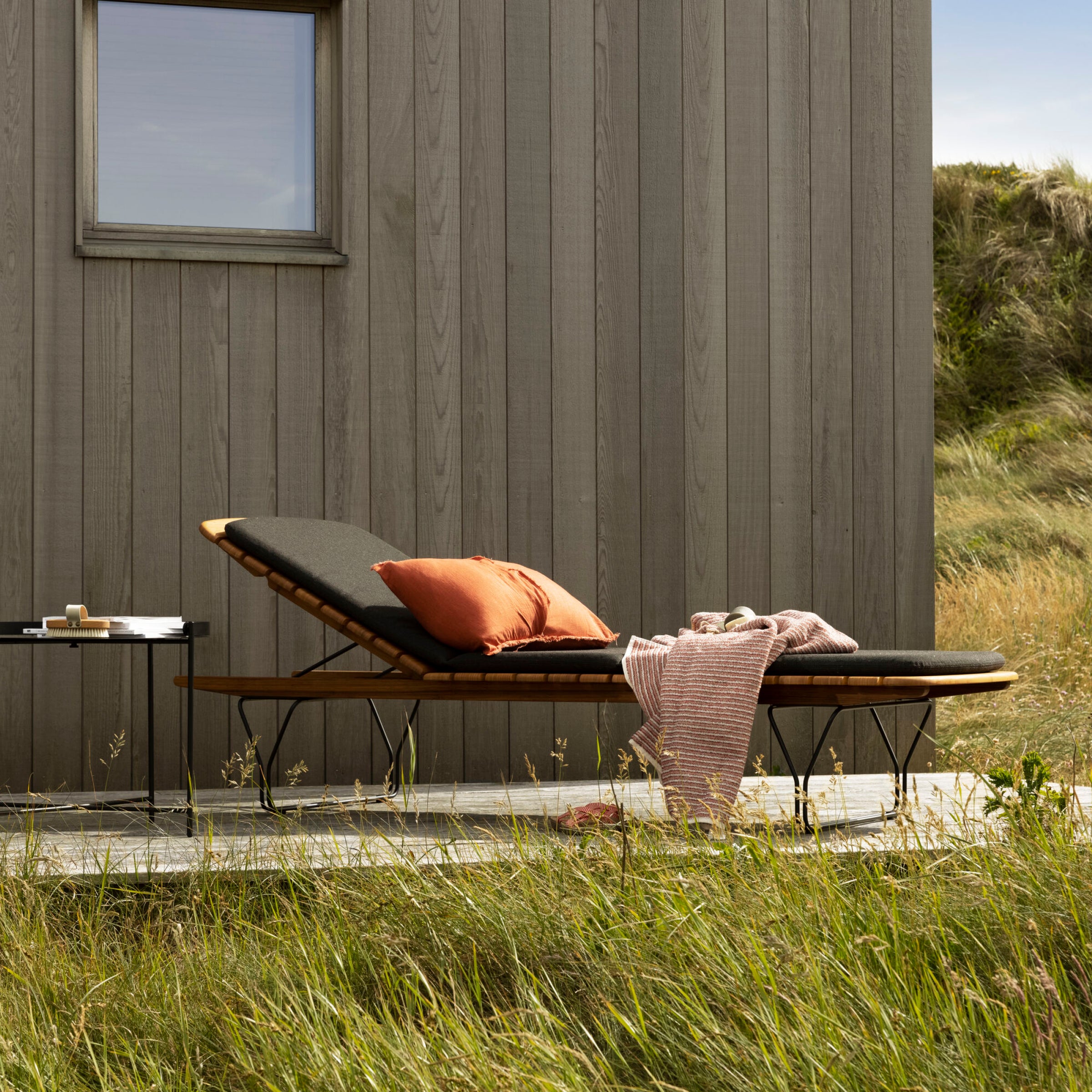A Houe Molo Sunbed with powder-coated black steel legs and bamboo lamellas boasts plush black cushions and two orange pillows. It features a pink striped blanket, next to a small table holding a plate and cup, set in tall grass near a wooden building.