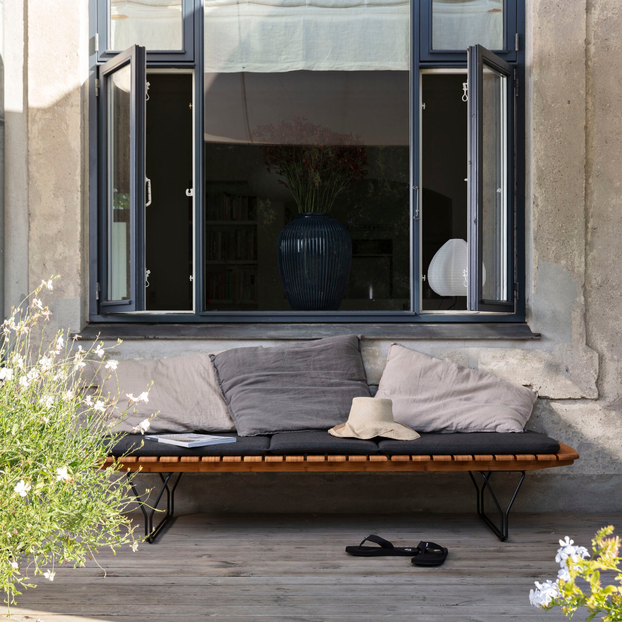 In front of a window, the MOLO Sunbed by Houe showcases gray cushions and a beige hat. A sandal rests on the bamboo-lamella-decked floor next to lush plants with white flowers, enhancing the natural appeal of the powder-coated black steel structure.