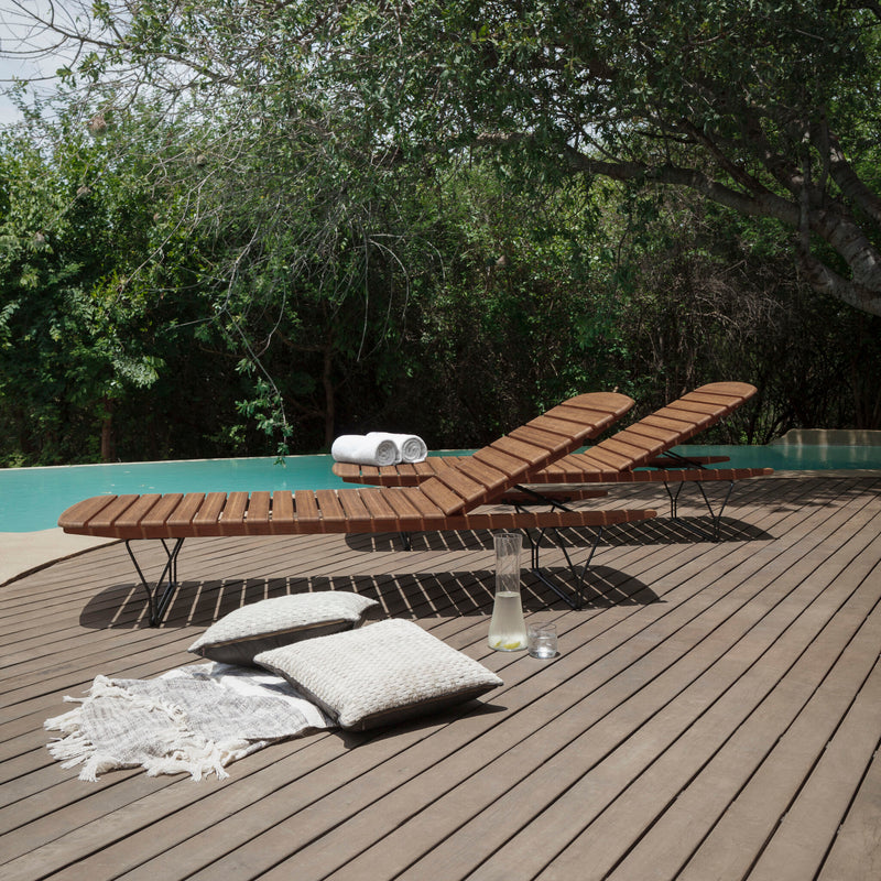 Two wooden lounge chairs with white towels next to a turquoise pool complement the Houe Molo Sunbed, crafted from powder-coated black steel. In the foreground, pillows and a beige throw rest on a deck near bamboo lamellas and a glass water pitcher. Lush green trees add serene tranquility.