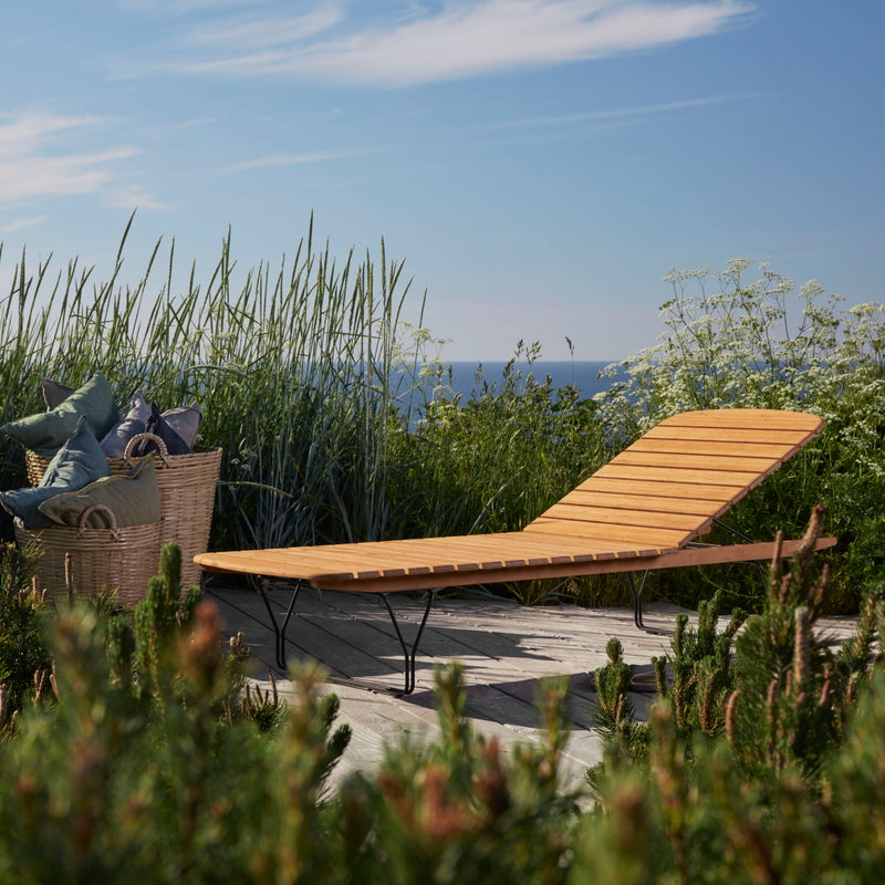 A wooden deck surrounded by lush greenery and tall grasses features a Houe Molo Sunbed and a wooden lounge chair. A basket with cushions adds to the serene atmosphere, while an ocean view under a clear blue sky completes the tranquil retreat.