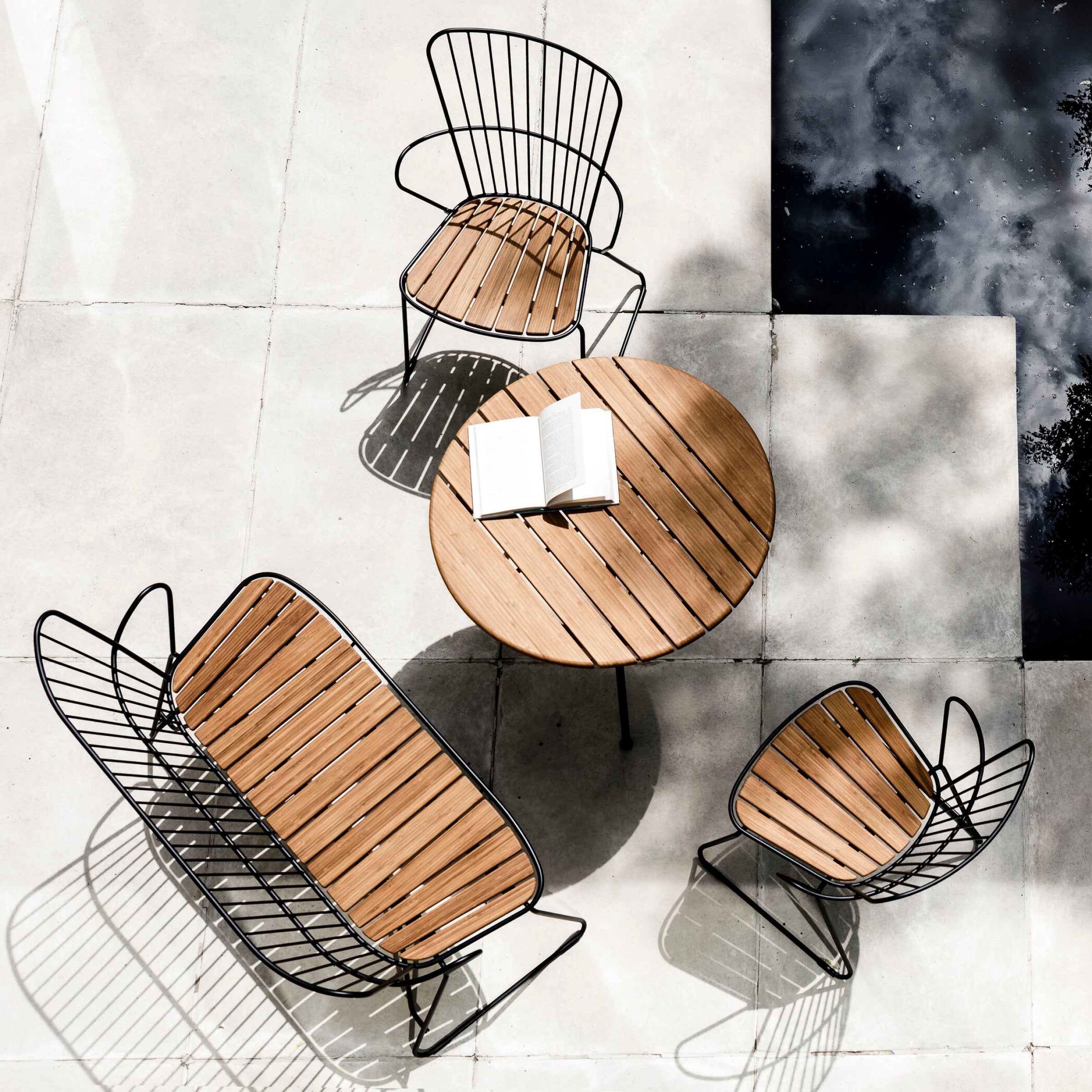 An aerial view displays a minimalist outdoor patio set on light tiles, featuring a round wooden table with an open book and three Houe Paon benches. Shadows and reflections from trees and a water feature highlight its Scandinavian design influence.