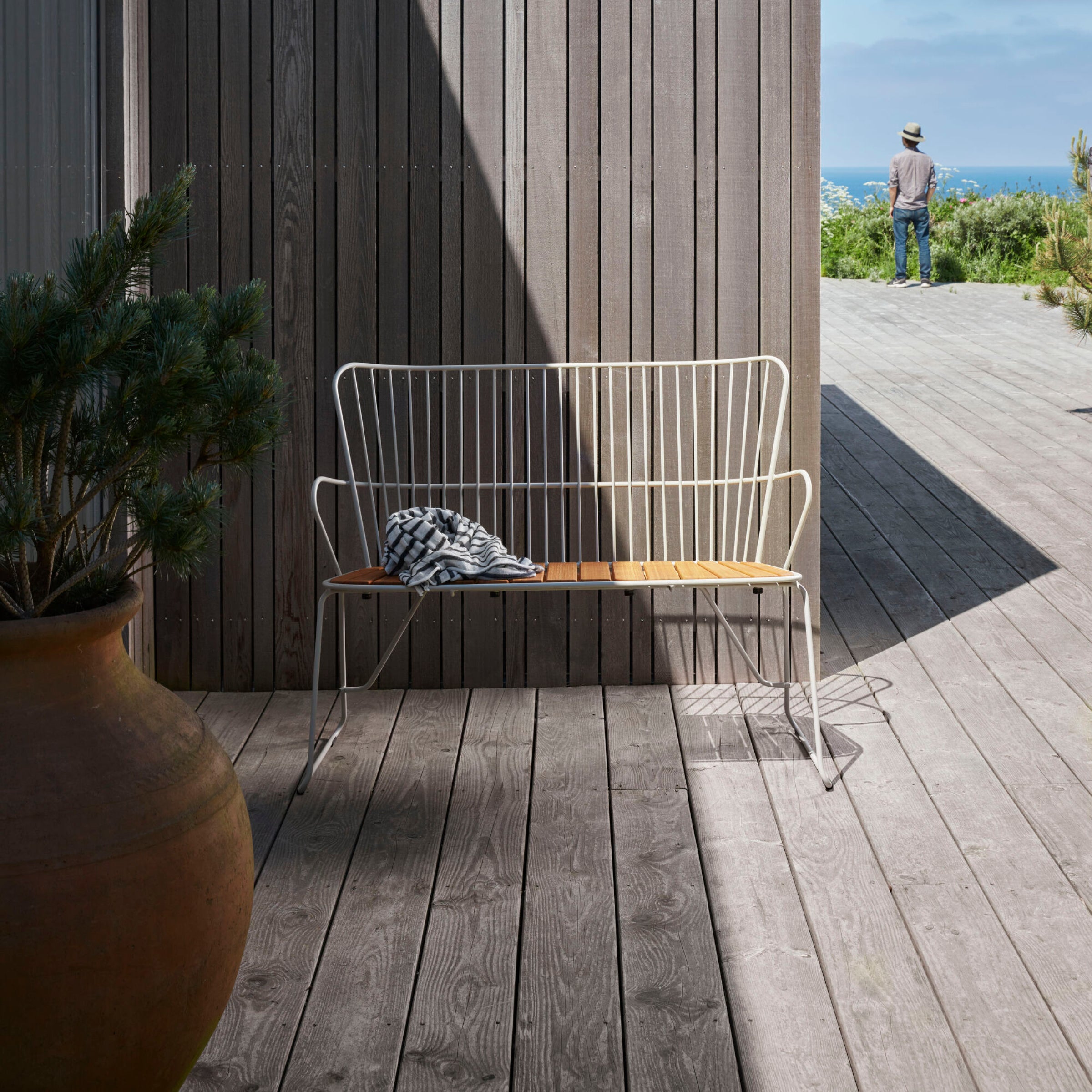 A Paon Bench by Houe, blending wood and metal in Scandinavian design, sits on the sunlit deck beside a large plant pot. A crumpled shirt lies on the bench as a hat-wearing person gazes at the ocean in the background.