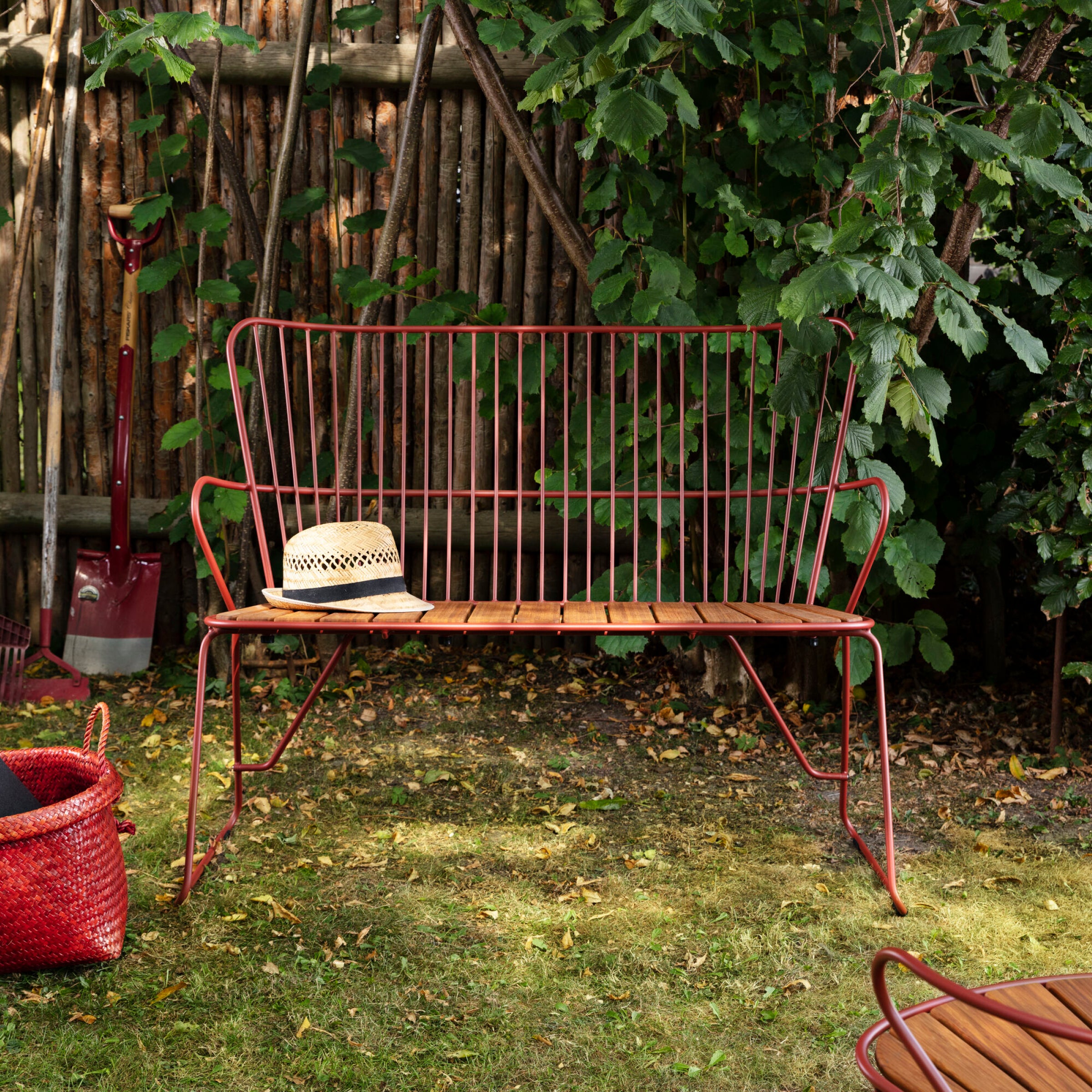 A PAON Bench by Houe, with a red metal frame and wooden seat, is on garden grass. A straw hat and book sit on the bench. Nearby are a red basket and shovel, with lush green plants against a wooden fence in the background.