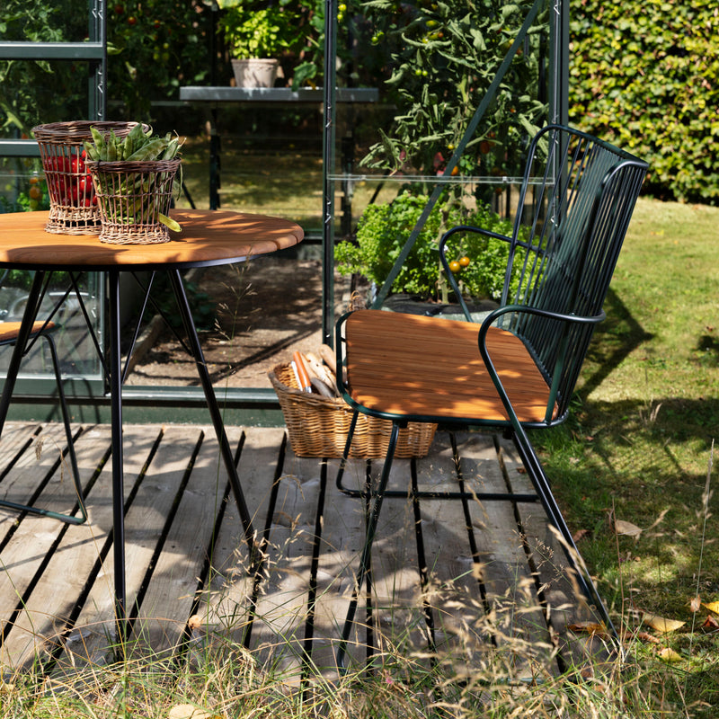A wooden patio set featuring a Houe Paon Bench surrounds a round table on a deck in the garden. A basket with rolled towels sits beside it. Greenery thrives through the glass structure, casting playful shadows as sunlight filters through, complementing the Scandinavian design ambiance.