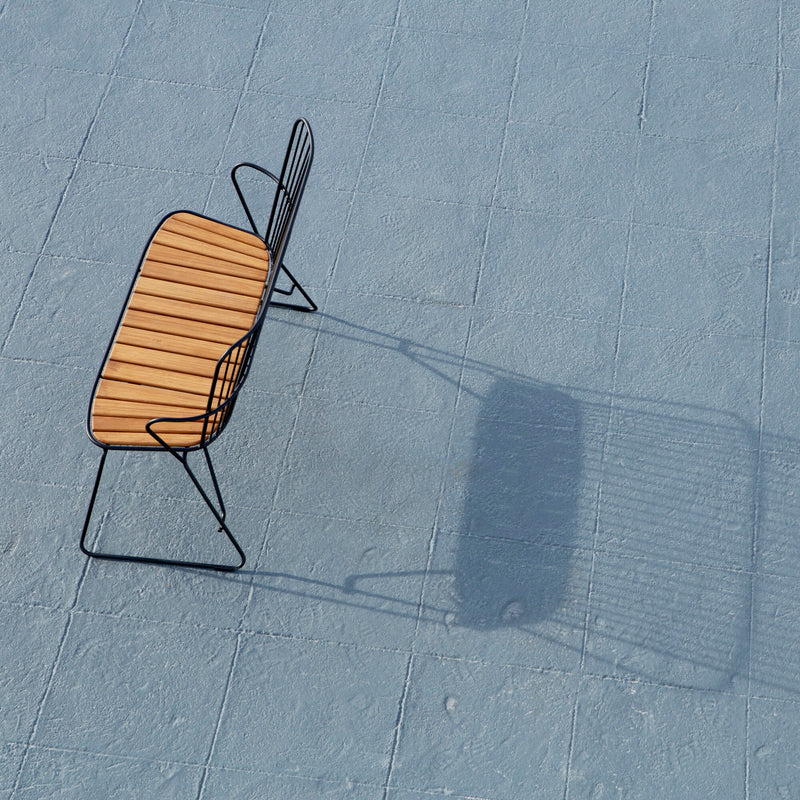 The Paon Bench by Houe features a slatted wooden seat with bamboo lamellas, Scandinavian-inspired design, and a sleek, black metal frame. Positioned on a light blue paved surface, it casts a distinct shadow suggestive of an open area under clear skies.
