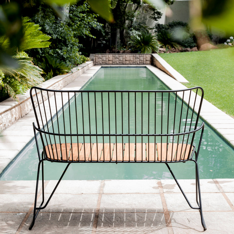 The Paon Bench by Houe, featuring a black metal frame and wooden seat, is placed on a stone patio overlooking a long narrow pool. Surrounded by lush greenery, the setup exudes tranquility and simplicity reminiscent of Scandinavian design.