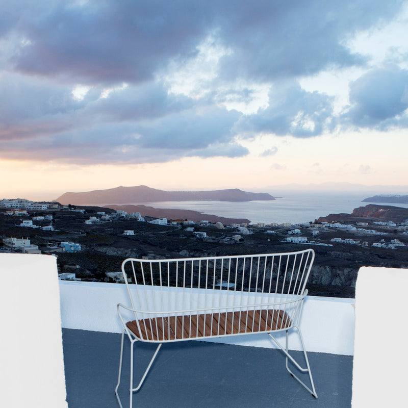 The Paon Bench by Houe, featuring a white metal frame with wooden slats in Scandinavian style, is perfectly placed on a terrace with views of an expansive landscape, including a distant town, rolling hills, and a cloudy sunset sky, creating a serene atmosphere.
