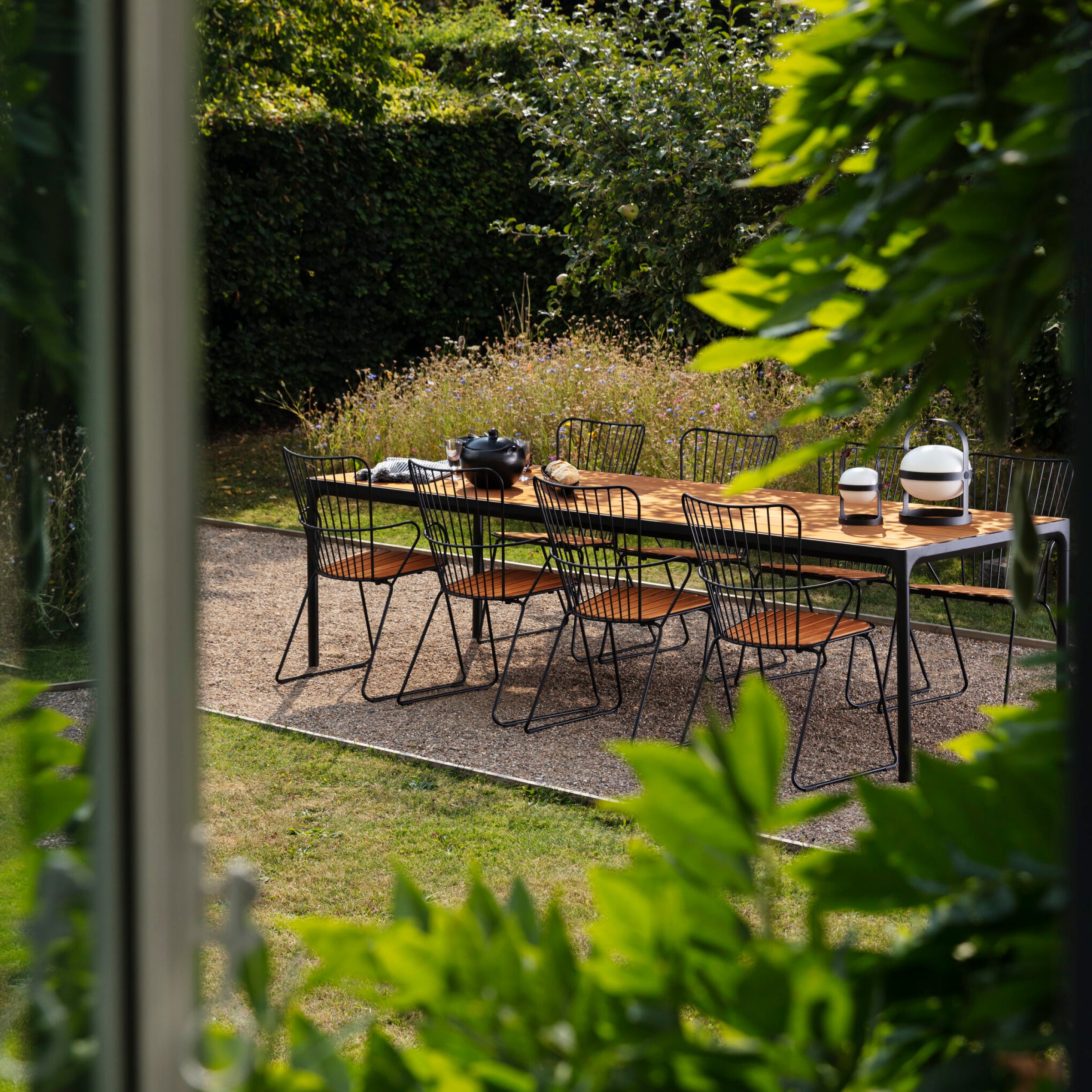 An outdoor dining area showcases Scandinavian style with a wooden table for eight, encircled by chic black Houe Paon Dining Chairs. Green foliage and lush shrubs frame the scene. On the table are a bowl and vase.