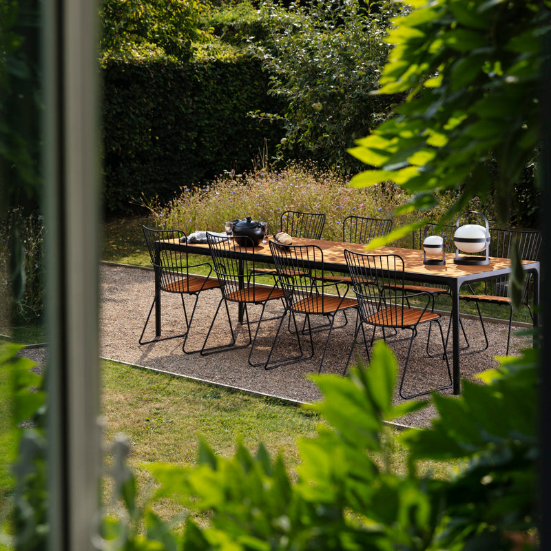 An outdoor dining area showcases Scandinavian style with a wooden table for eight, encircled by chic black Houe Paon Dining Chairs. Green foliage and lush shrubs frame the scene. On the table are a bowl and vase.