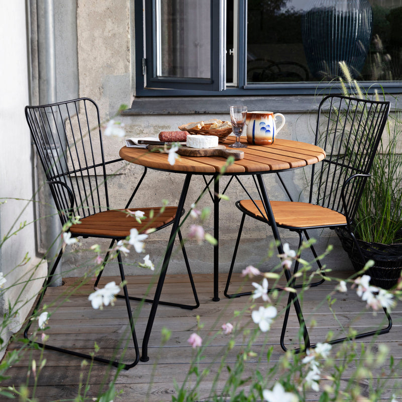 A cozy outdoor patio features a round wooden table and two Houe Paon Dining Chairs. The table is adorned with a teapot, food plate, glass, and mug. Delicate white flowers are in the foreground, with the window reflecting greenery echoing Scandinavian design.
