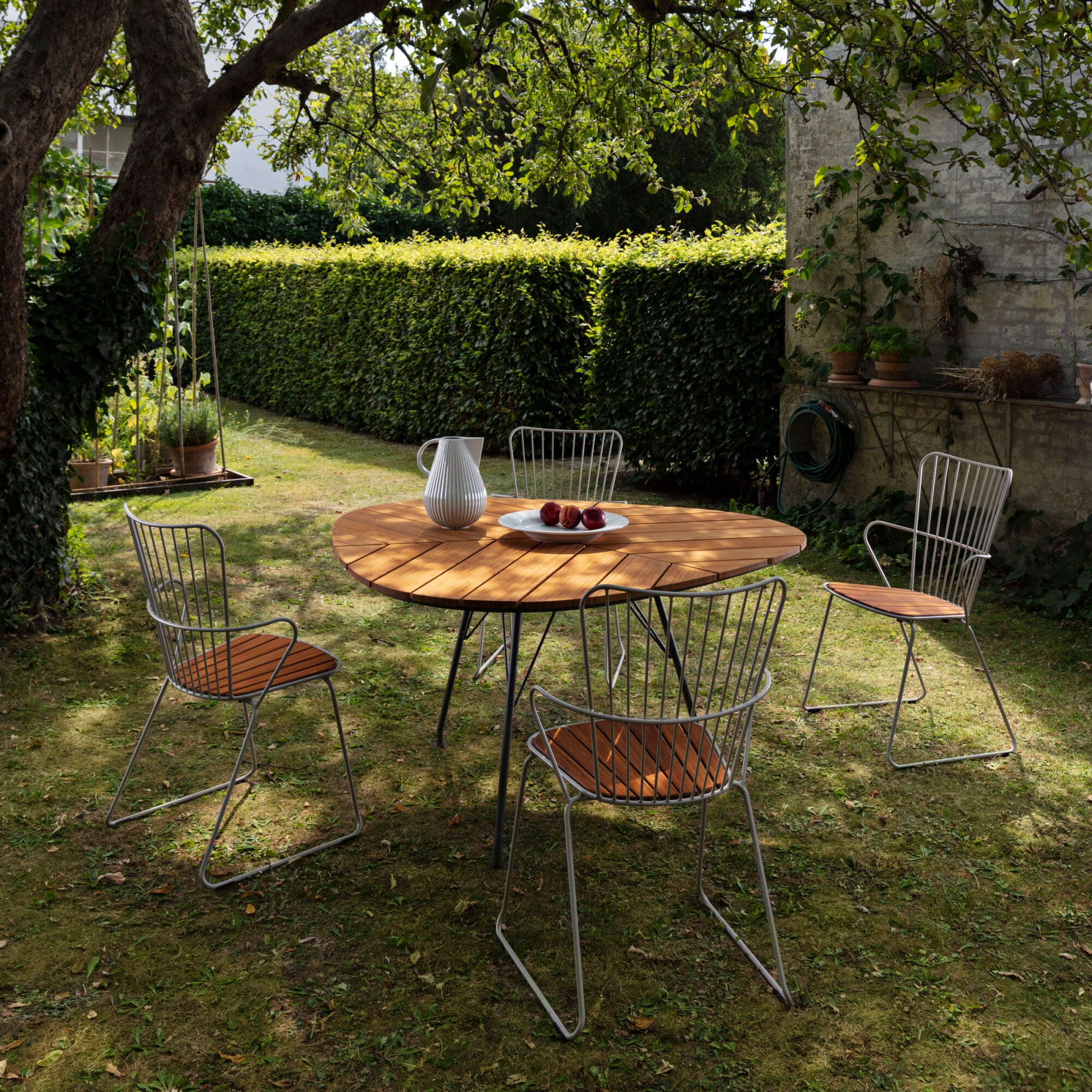 In a garden, a round wooden table adorned with a white pitcher and bowl of apples is encircled by four Houe Paon Dining Chairs. Shaded by a tree, it exudes Scandinavian elegance. A stone wall and hedge frame the scene, enhancing the serene outdoor ambiance.