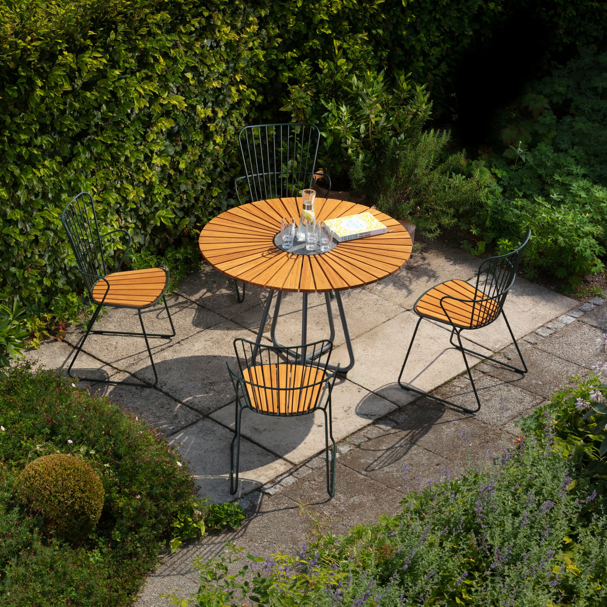 On a stone patio, a round wooden table is paired with four Houe Paon Dining Chairs, surrounded by lush greenery. A glass of water and a magazine rest on the table, encapsulating the peaceful Scandinavian design essence in this serene outdoor setting.