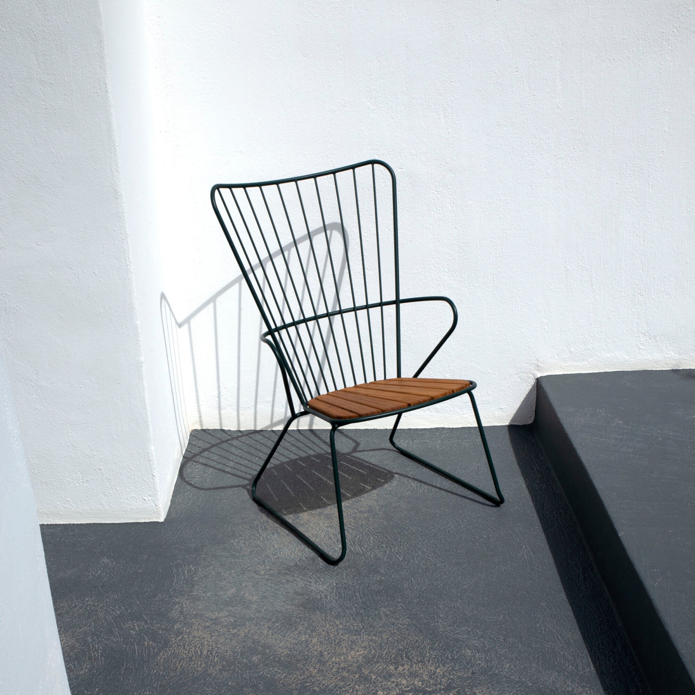 The Paon Lounge Chair by Houe, featuring a wooden seat and powder-coated metal frame, is set on a grey concrete floor. Positioned in the corner of two white walls, it casts a shadow under the sunlight.