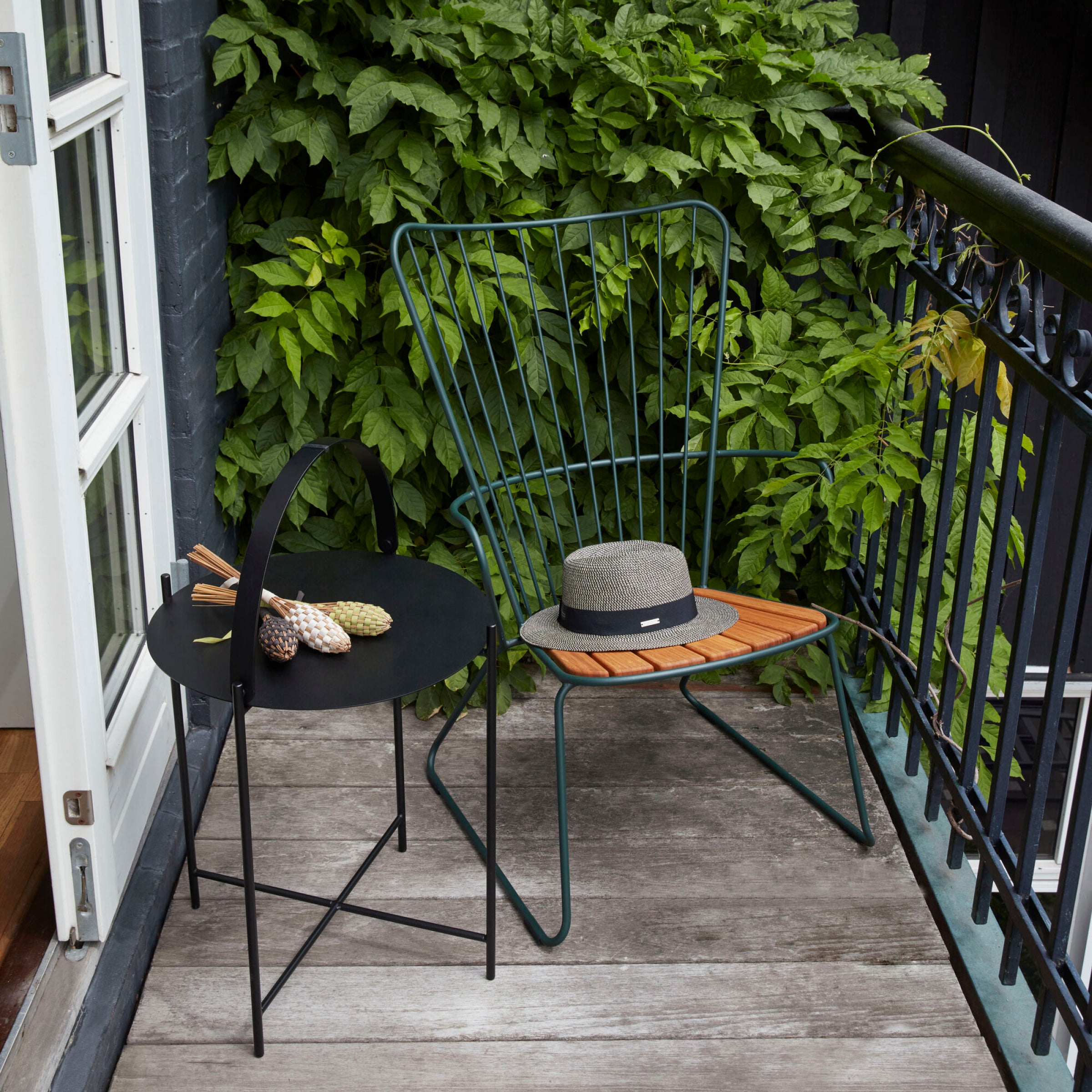 On a small balcony, a black side table holds two pineapples by a powder-coated steel railing. A Houe Paon Lounge Chair with bamboo lamellas and a wooden seat has a gray hat on it, as lush greenery climbs the background wall.