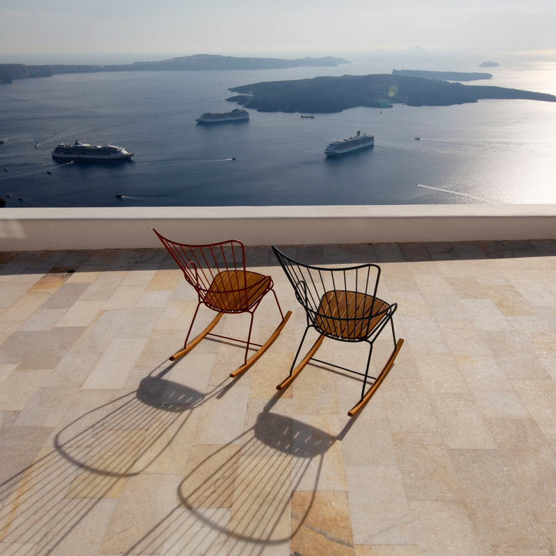 Two elegant Houe Paon Rocking Chairs grace a stone patio overlooking a serene sea and islands. Cruise ships and boats glide across the water under a clear sky, their reflections offering a peaceful, picturesque coastal view.